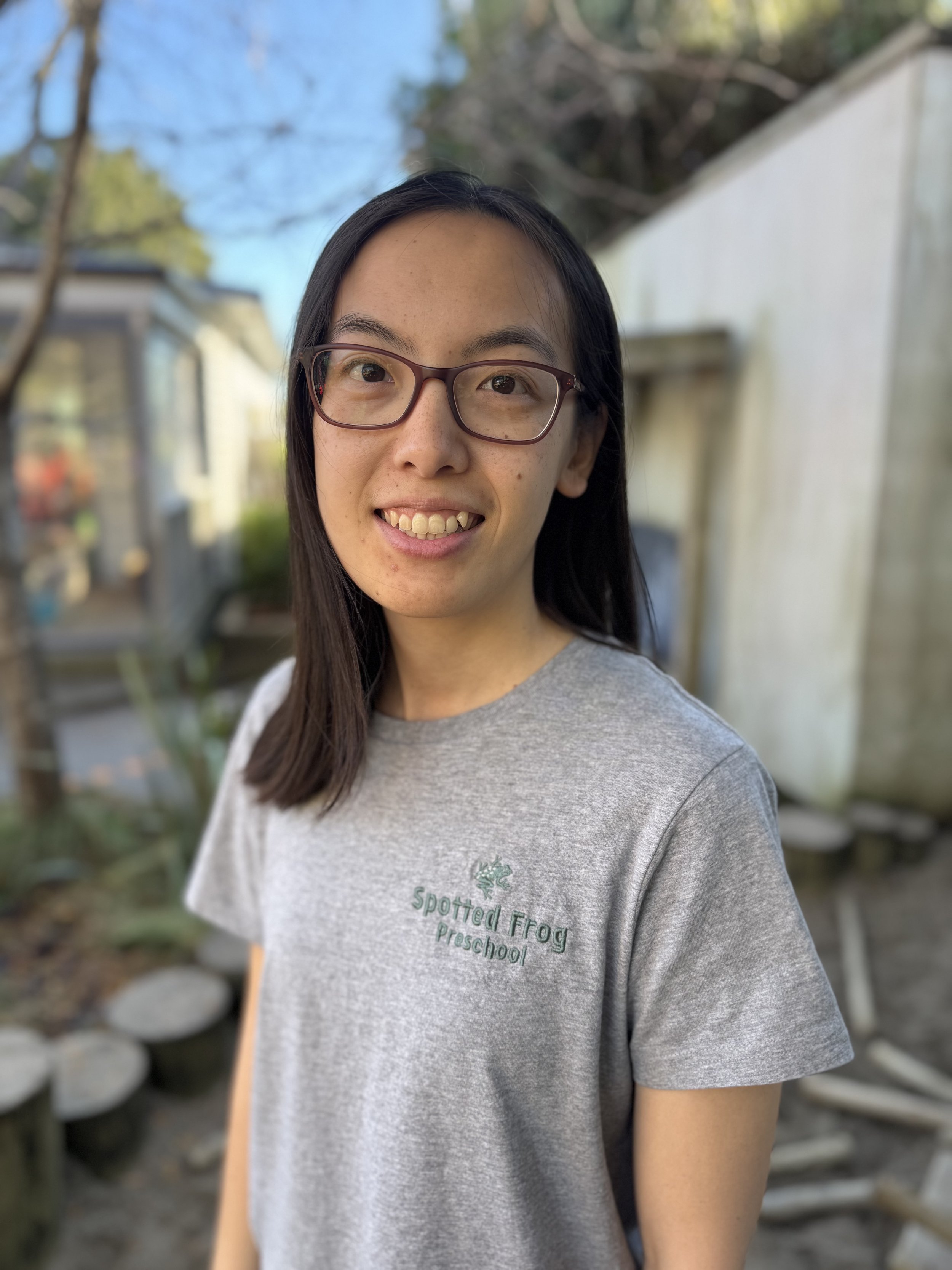 A female ECE teacher smiling and standing outdoors with a blurred garden background, wearing a grey t-shirt with the words "Spotted Frog Preschool" and a frog logo printed on it.