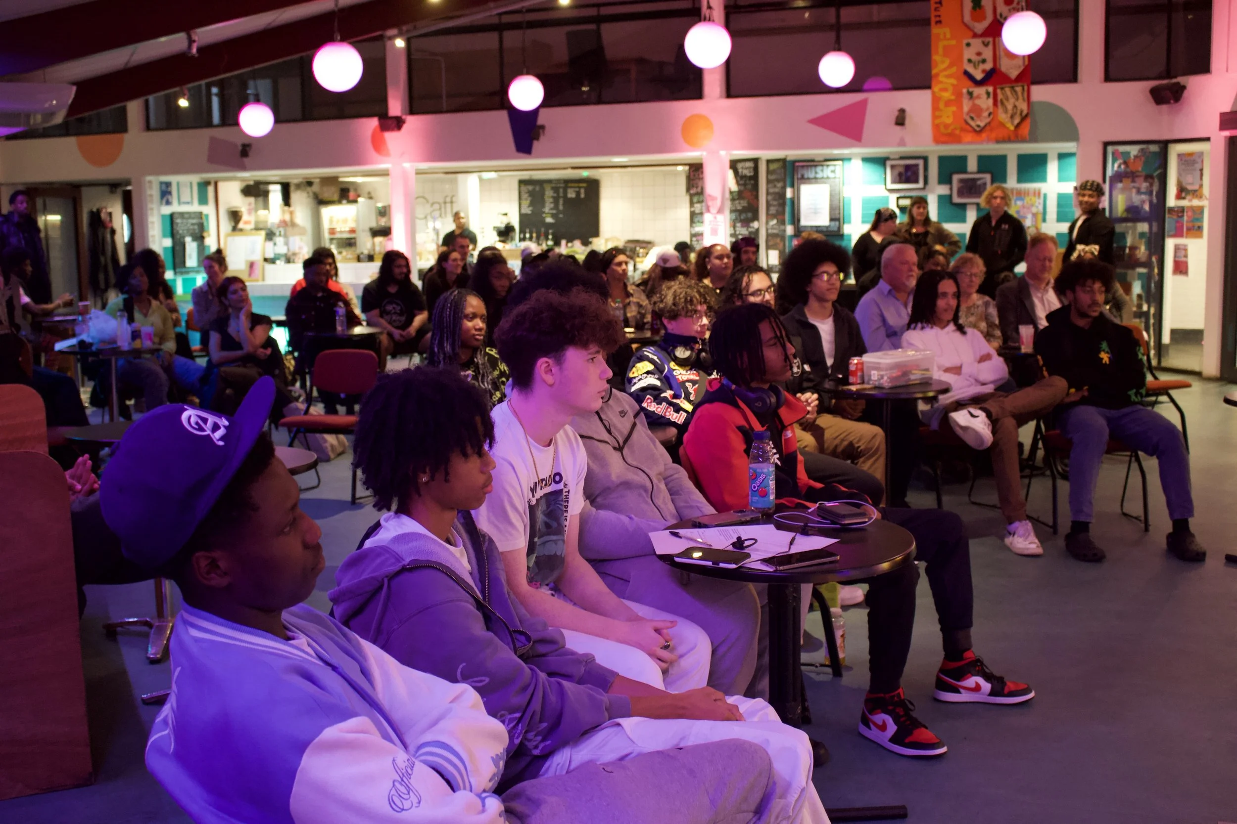 A diverse group of people seated in a cafe-like setting, attentively watching a performance or presentation. The atmosphere is casual, with colorful lighting and decor.