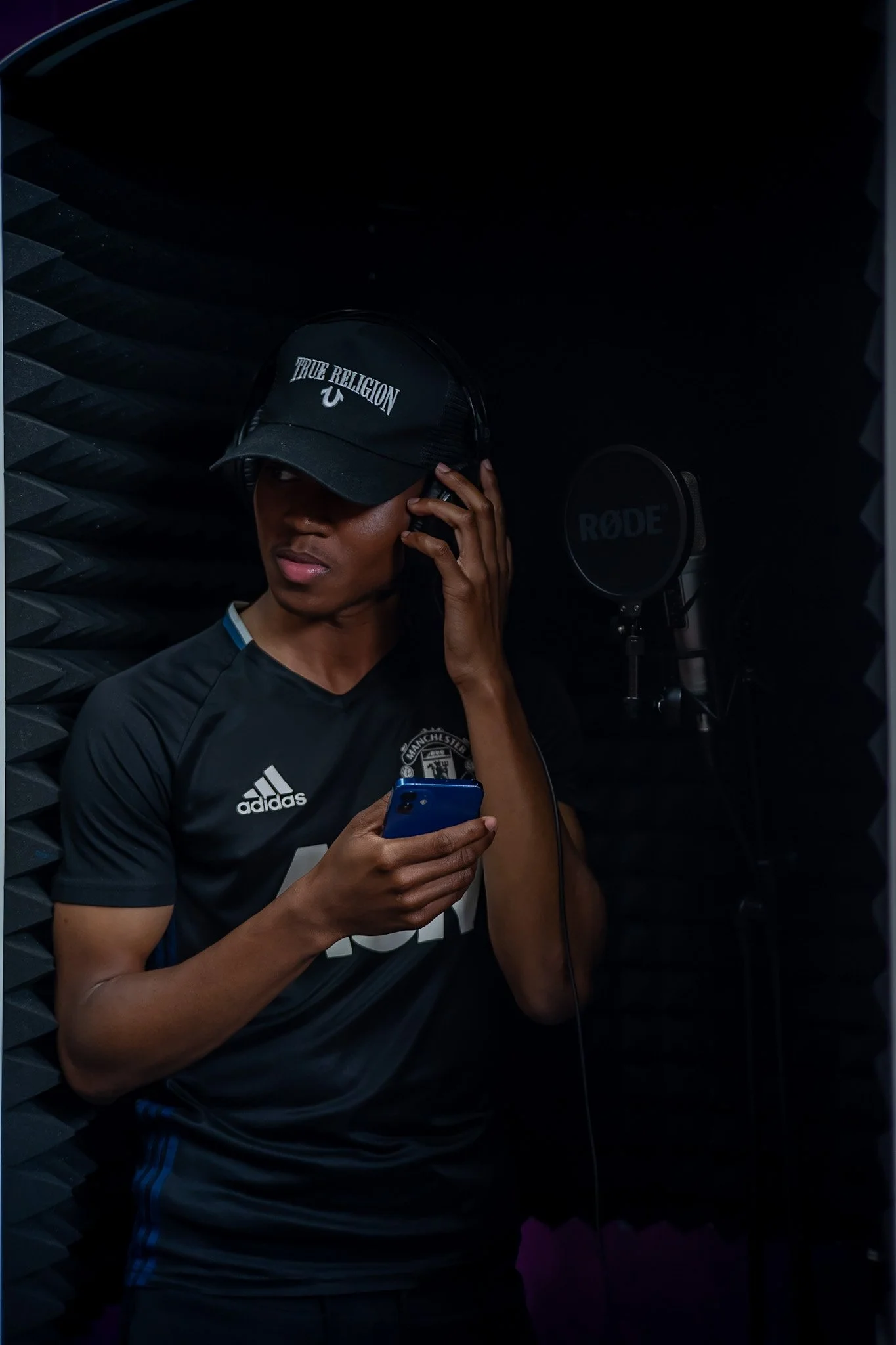 A young man in a black Manchester United jersey and a 'True Religion' cap in a recording booth, wearing headphones and standing beside a Rode microphone, holding a smartphone.