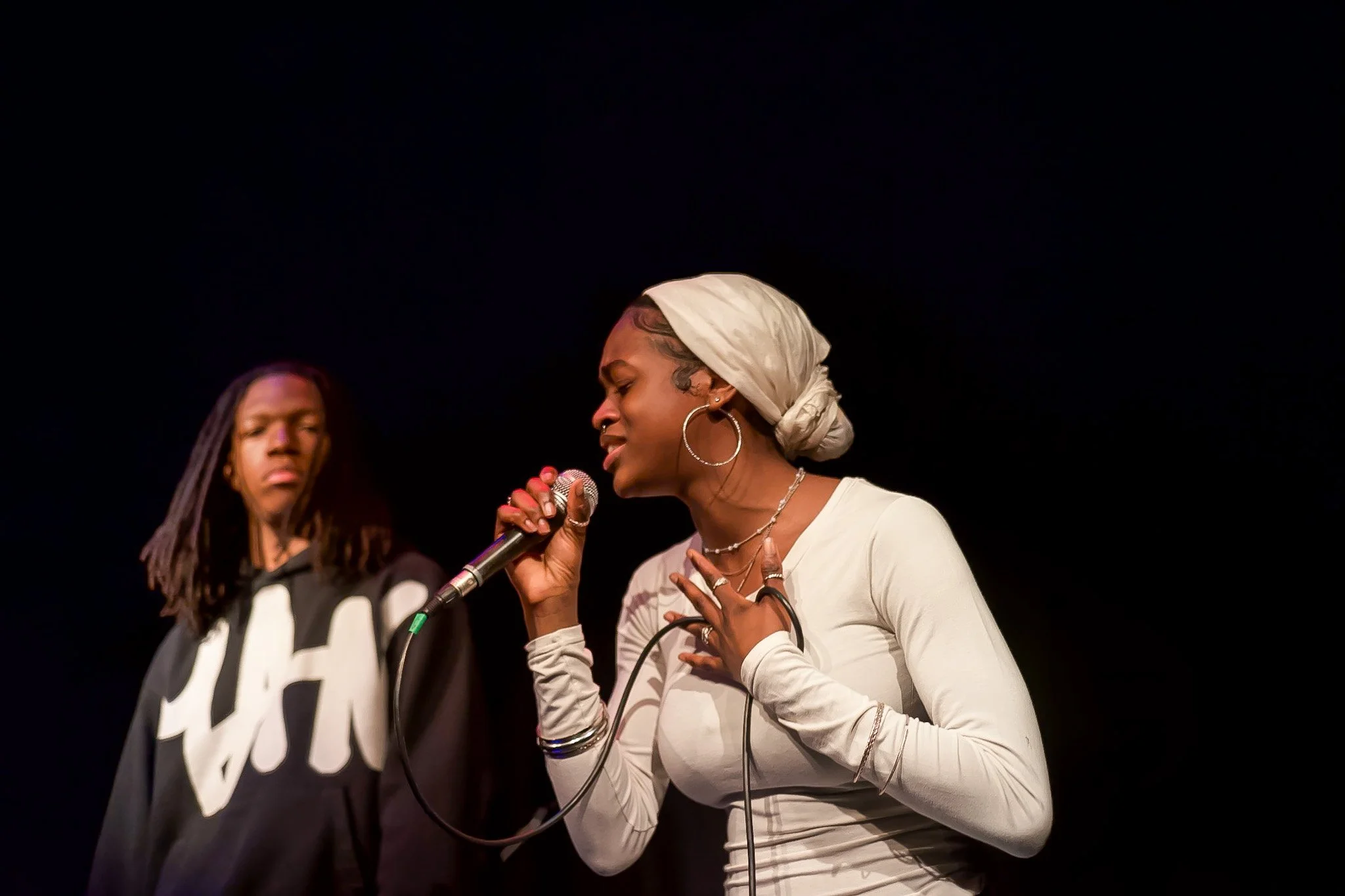 A female singer holding a microphone, performing passionately on stage with a male musician in the background.
