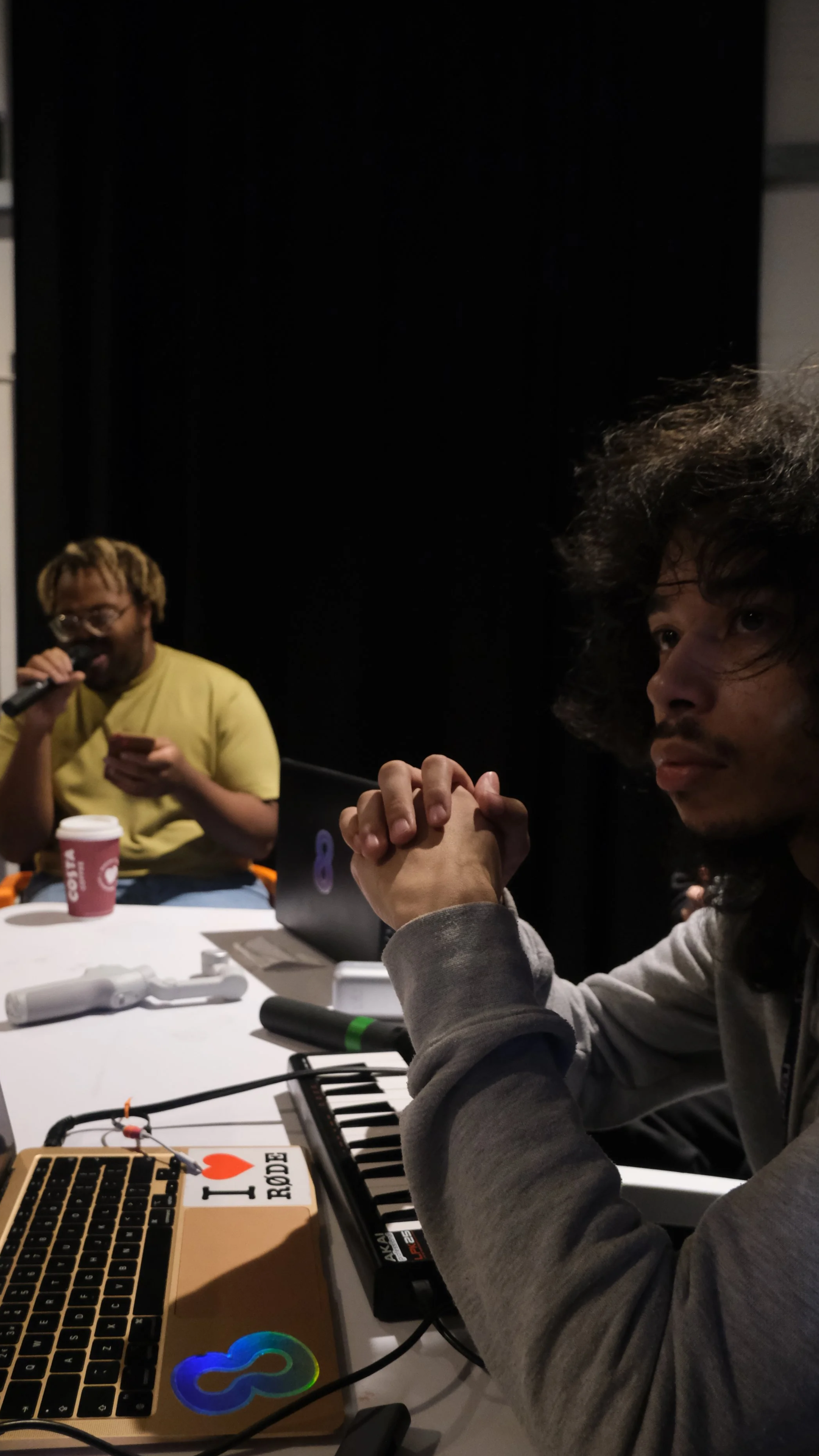 Two young men sitting at a table with music equipment inside a dark room. One is wearing a yellow shirt and glasses, holding a microphone and a phone; the other has curly hair and a hoodie, with hands clasped on the table.