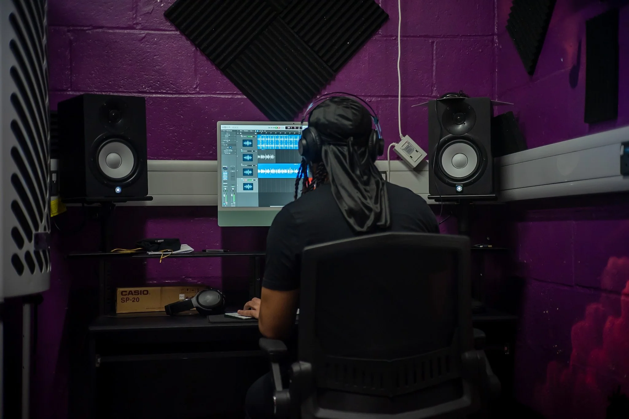 Person with headphones working at a music production computer in a purple-walled recording studio with speakers and acoustic foam.