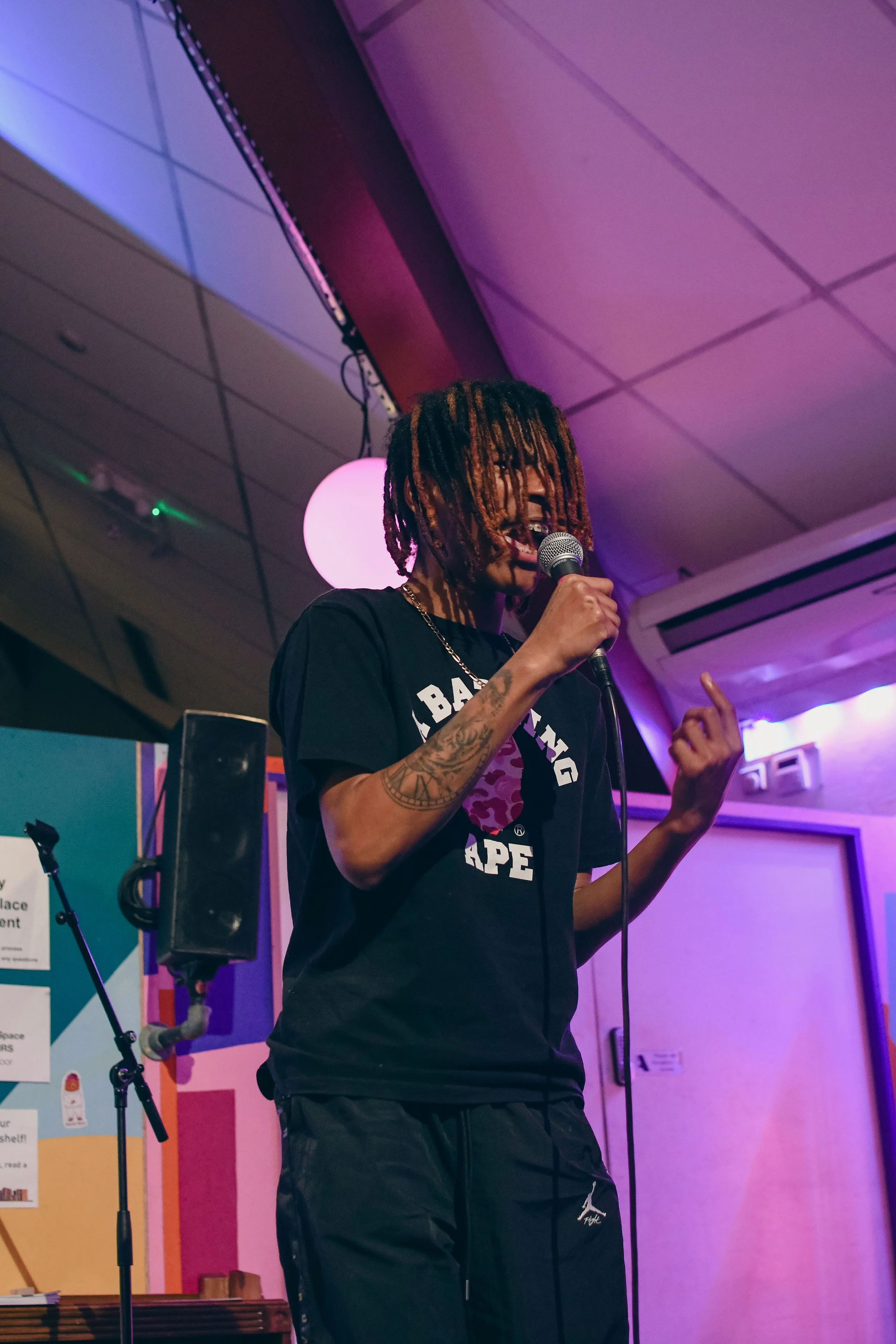 Person performing on stage with a microphone, wearing a black BAPE t-shirt, under colorful lighting.