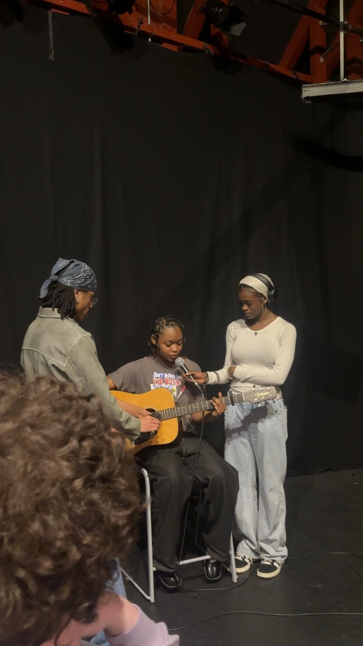 Three people in a music practice or performance setting, one seated playing a guitar, one holding a microphone, and another standing near them, in front of a black backdrop.