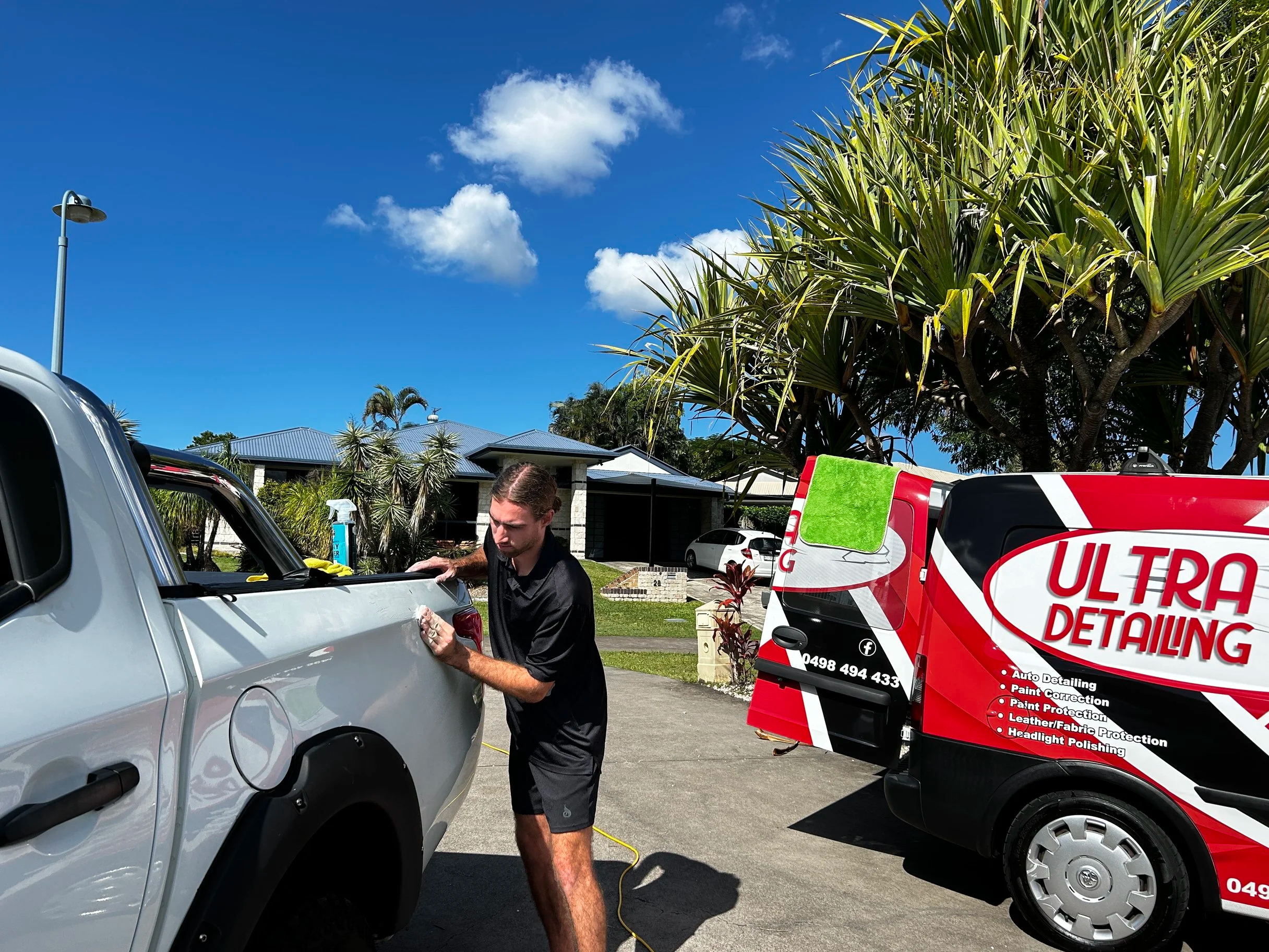 Man working on a white dual cab ute outside with a red and white ultra detail cleaning van nearby, under a blue sky with puffy white clouds, surrounded by green trees.