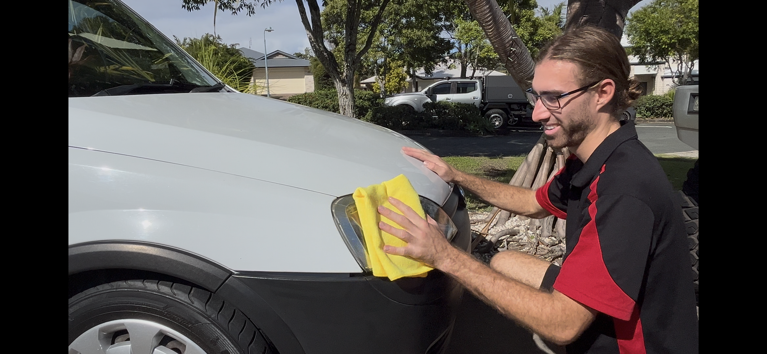 A man wearing glasses and a black and red shirt is cleaning the front of a white vehicle with a yellow microfibre cloth.