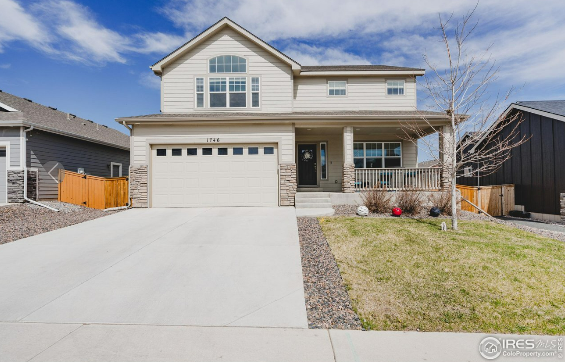 Front view of a two-story house with a concrete driveway, beige siding, stone accents, a small front porch, and a leafless tree in the yard.