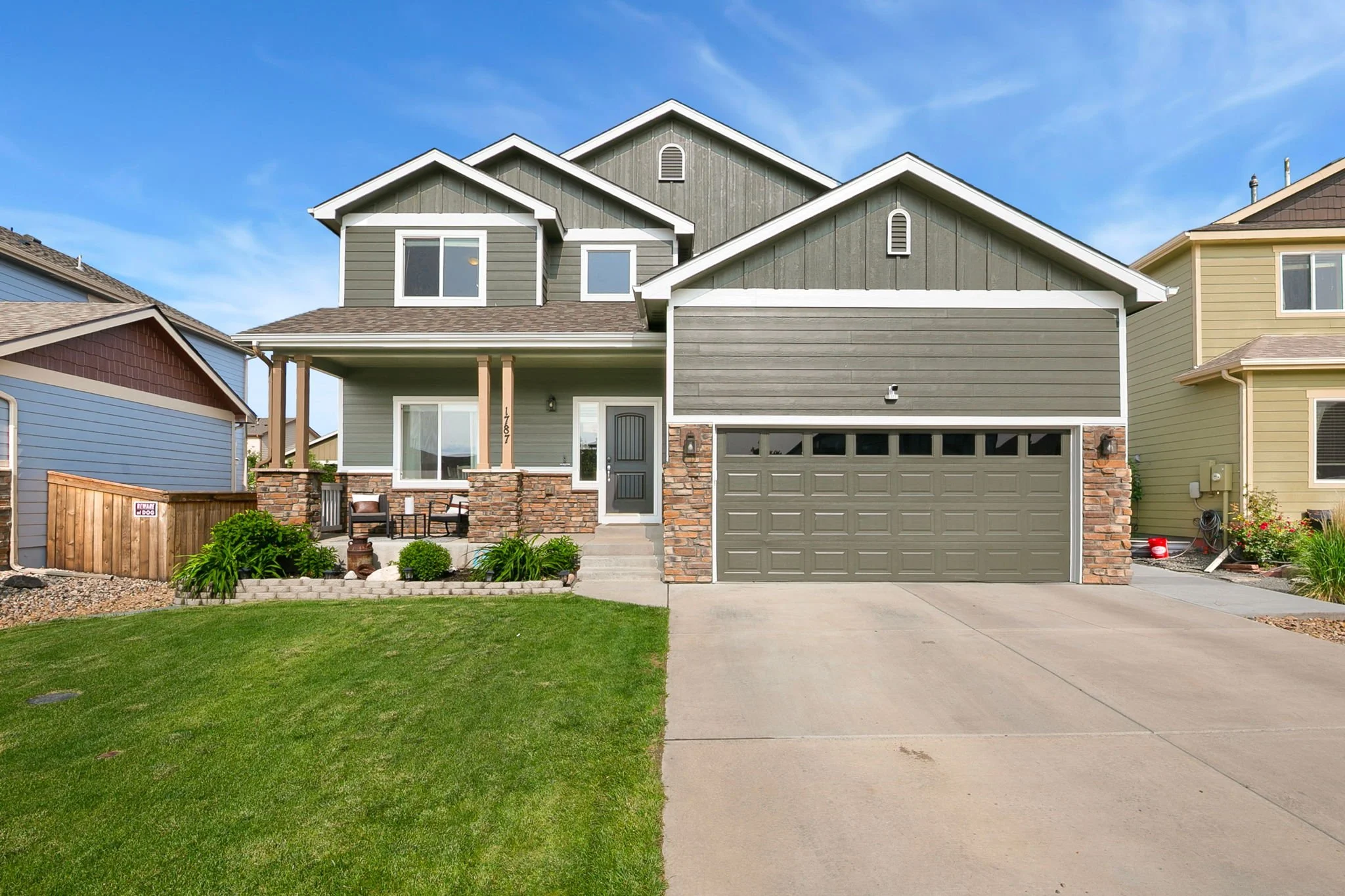 Modern two-story house with gray siding, stone accents, a double garage, front porch with seating, and a well-maintained lawn under a clear blue sky.