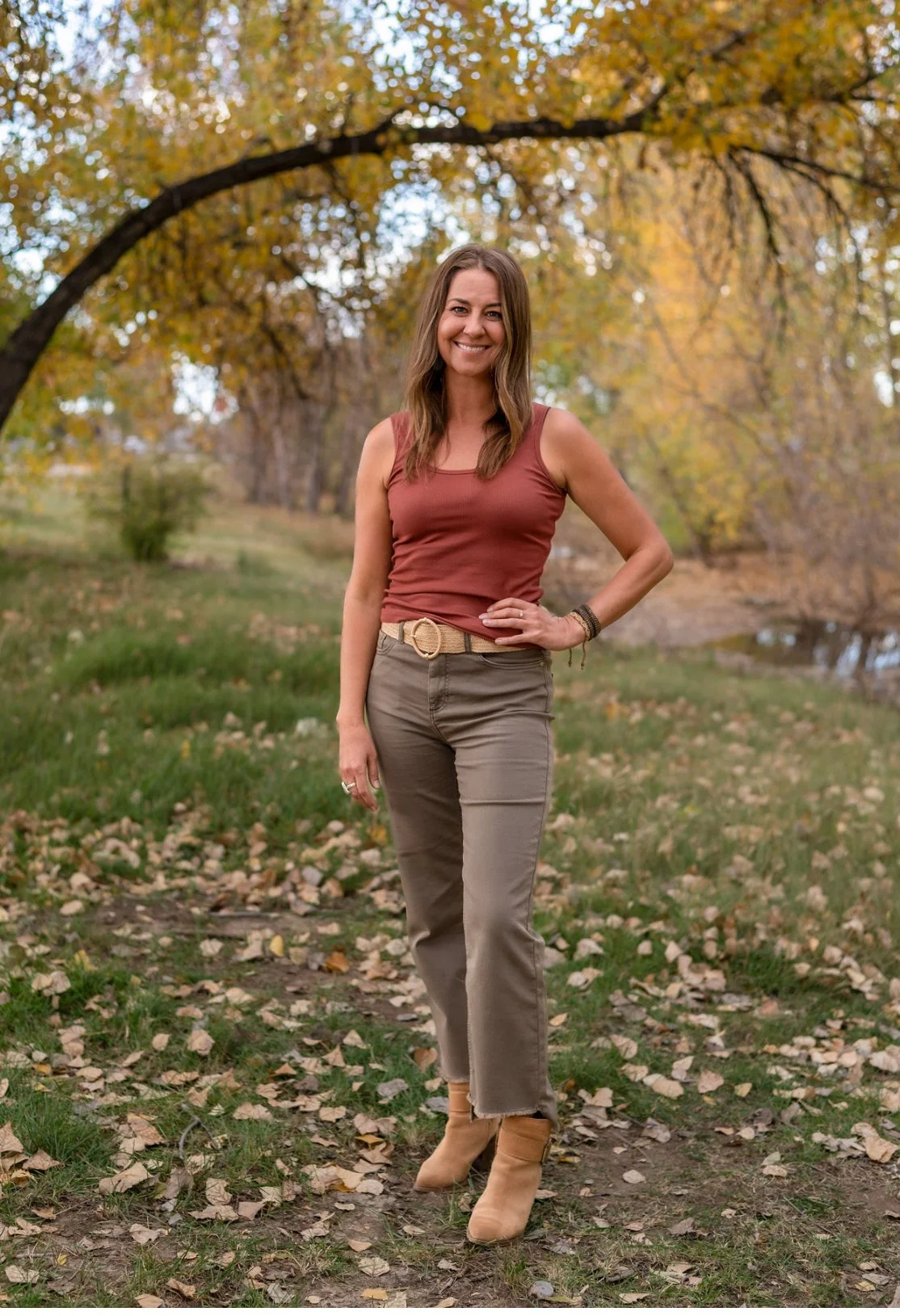 A woman standing outdoors in a park during fall, surrounded by yellow and orange leaves, wearing a rusty red sleeveless top, beige pants, and tan ankle boots, smiling at the camera.