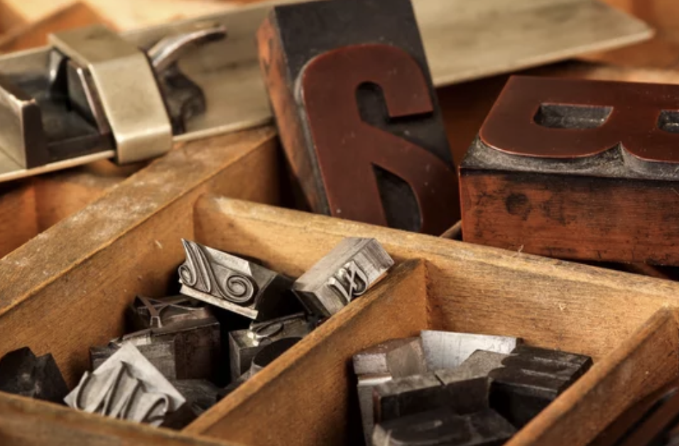 Close-up of a wooden box containing various vintage metal letterpress printing blocks, some showing decorative designs and others with letters.