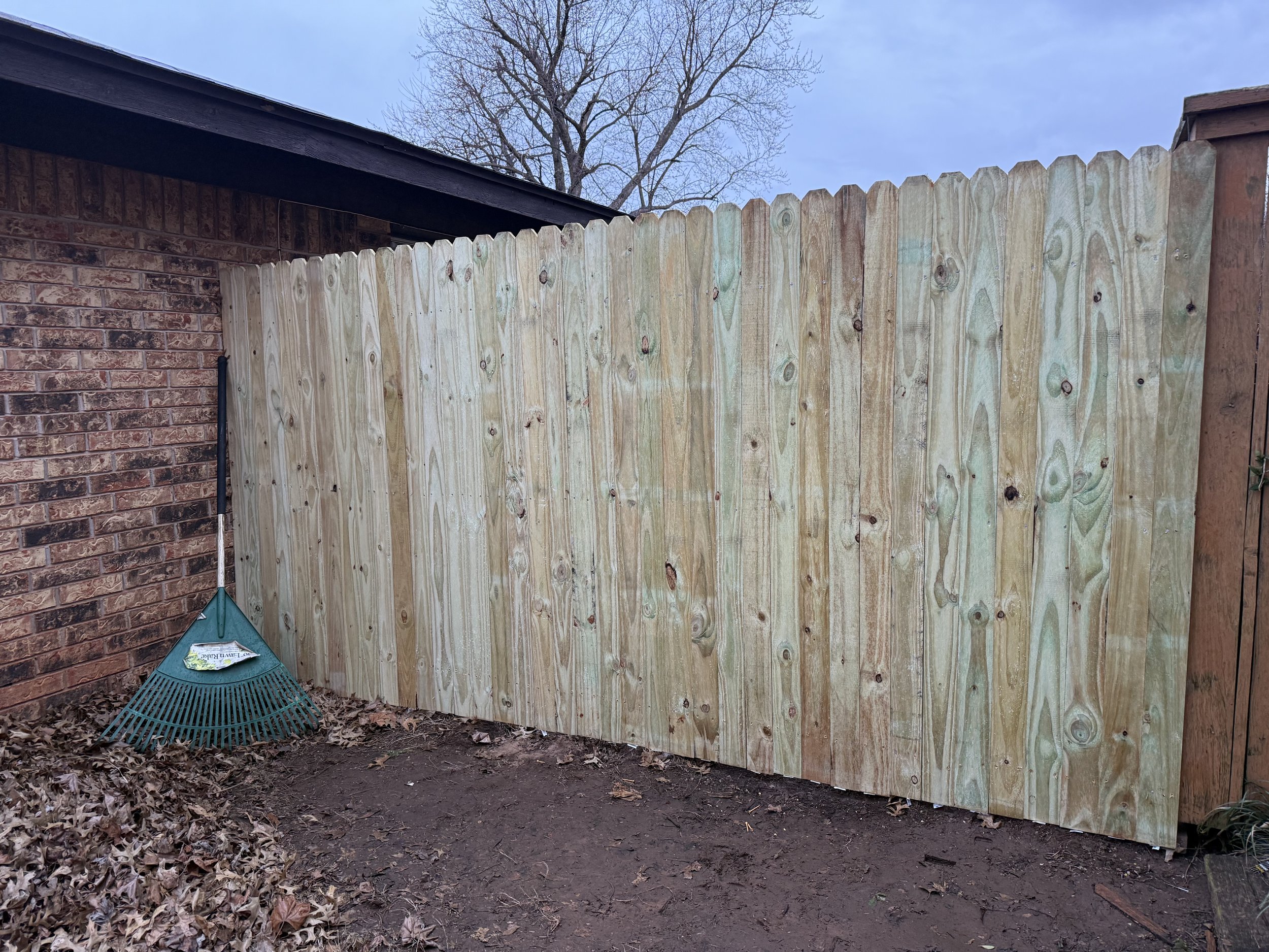 A wooden fence next to a brick wall with a green rake leaning against it. The ground is covered with dry leaves and dirt.