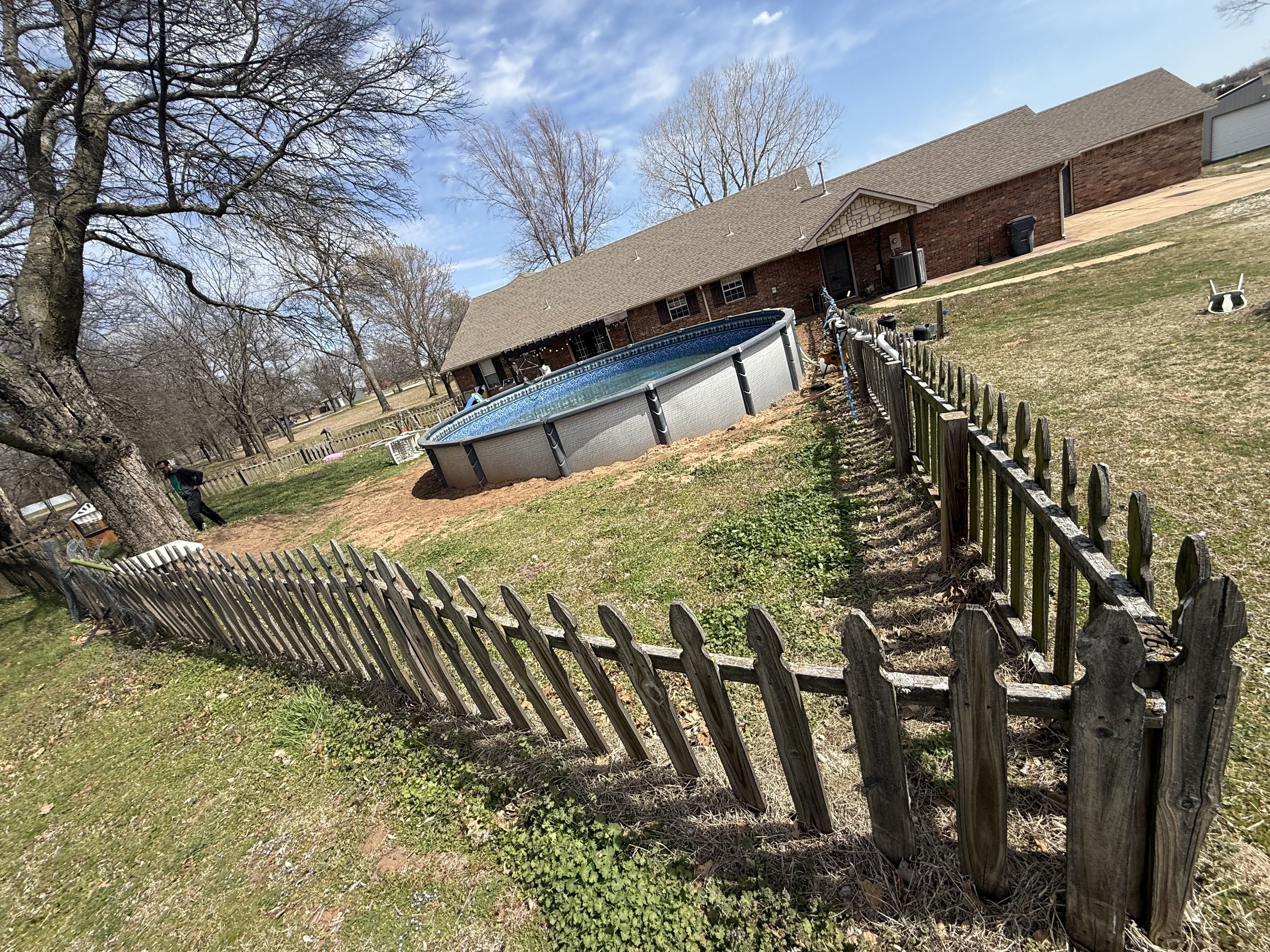 Backyard scene with an above-ground pool, wooden fence, brick house, and trees during early spring.