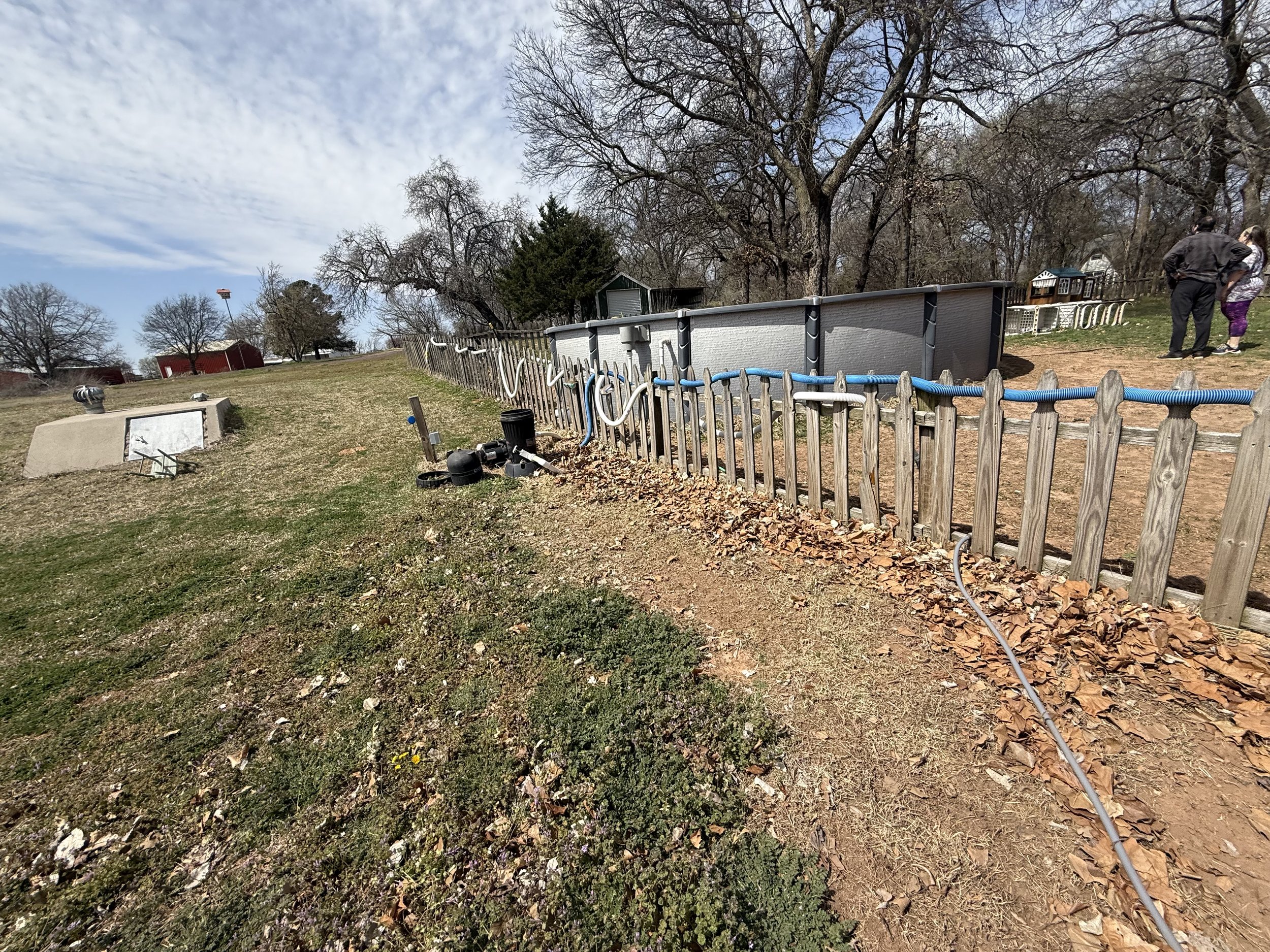 Outdoor scene with an above-ground pool, wooden fence, scattered leaves, grassy area, two people standing, and trees in the background.