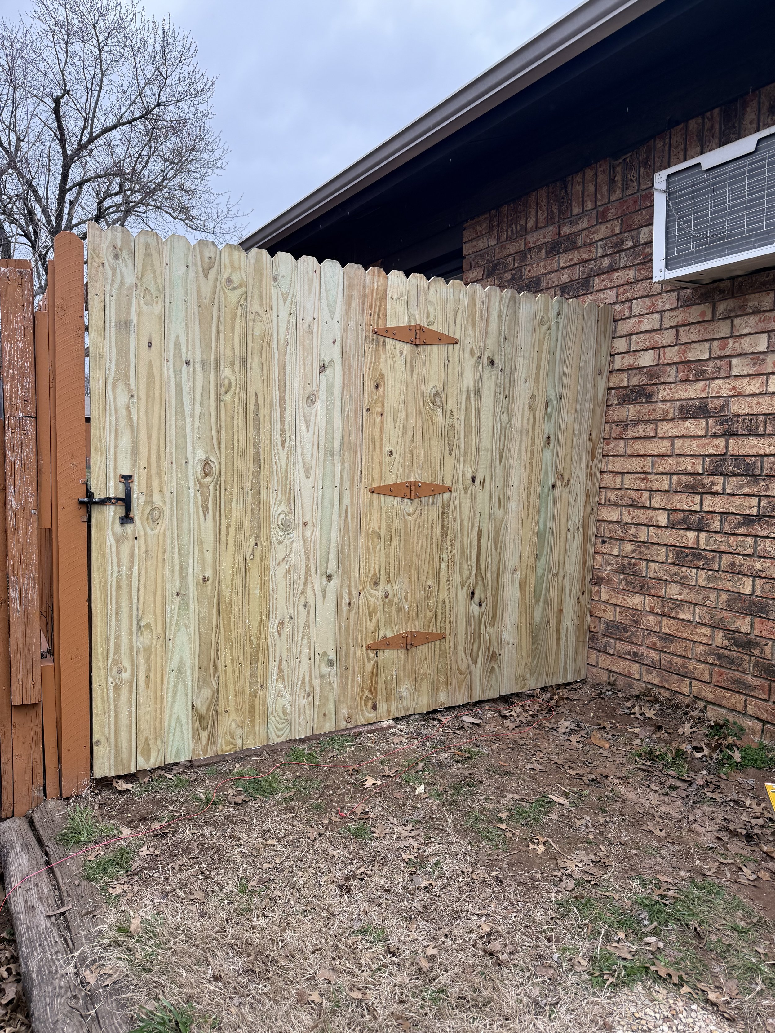 Wooden gate next to a brick wall of a house, with an air conditioning unit above. The foreground features dry grass and some leaves, while a tree with bare branches is visible in the background.
