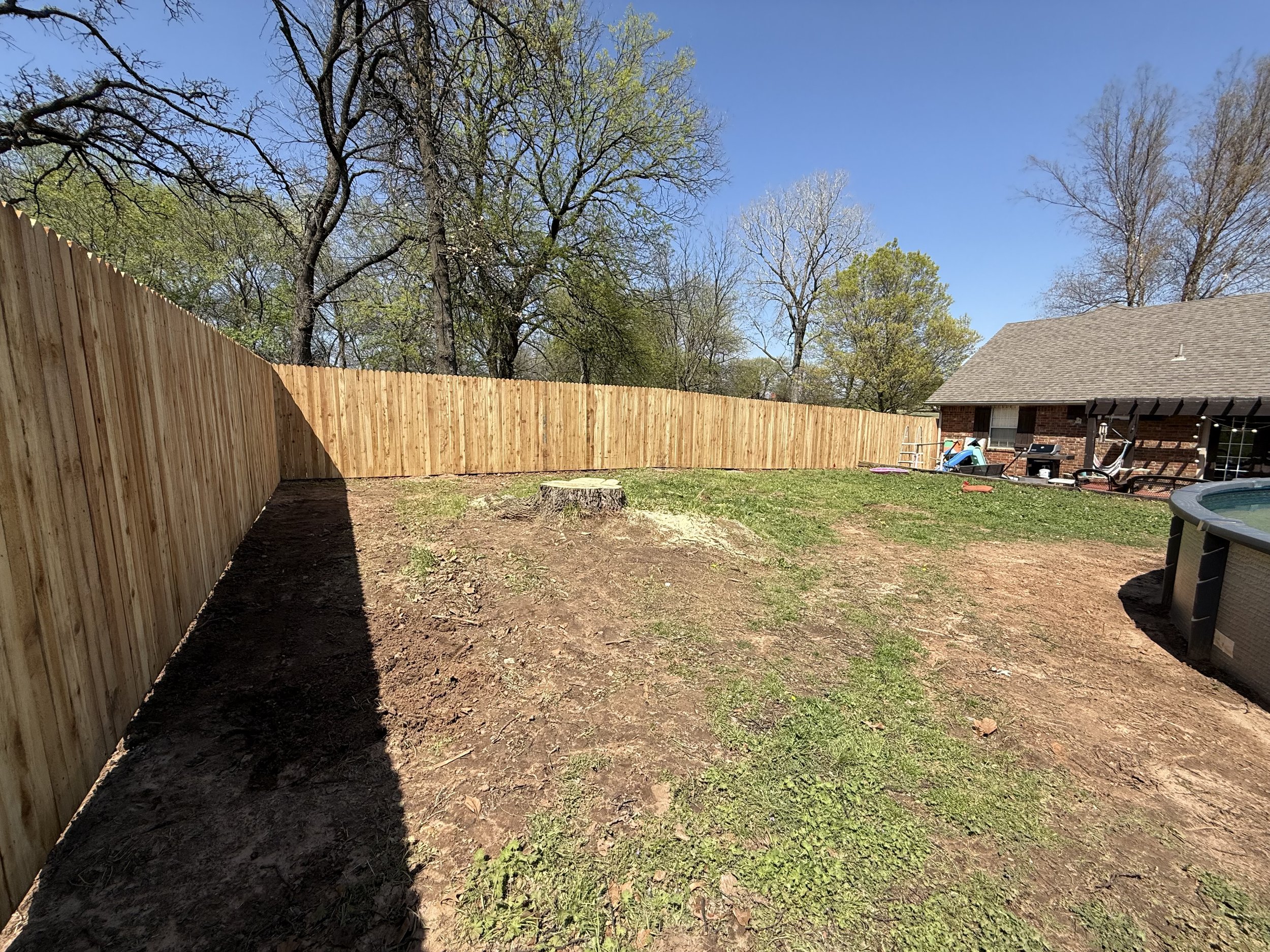 Backyard with new wooden privacy fence, surrounding a grassy area with some bare patches, a tree stump, an above-ground pool, and a house with patio furniture.