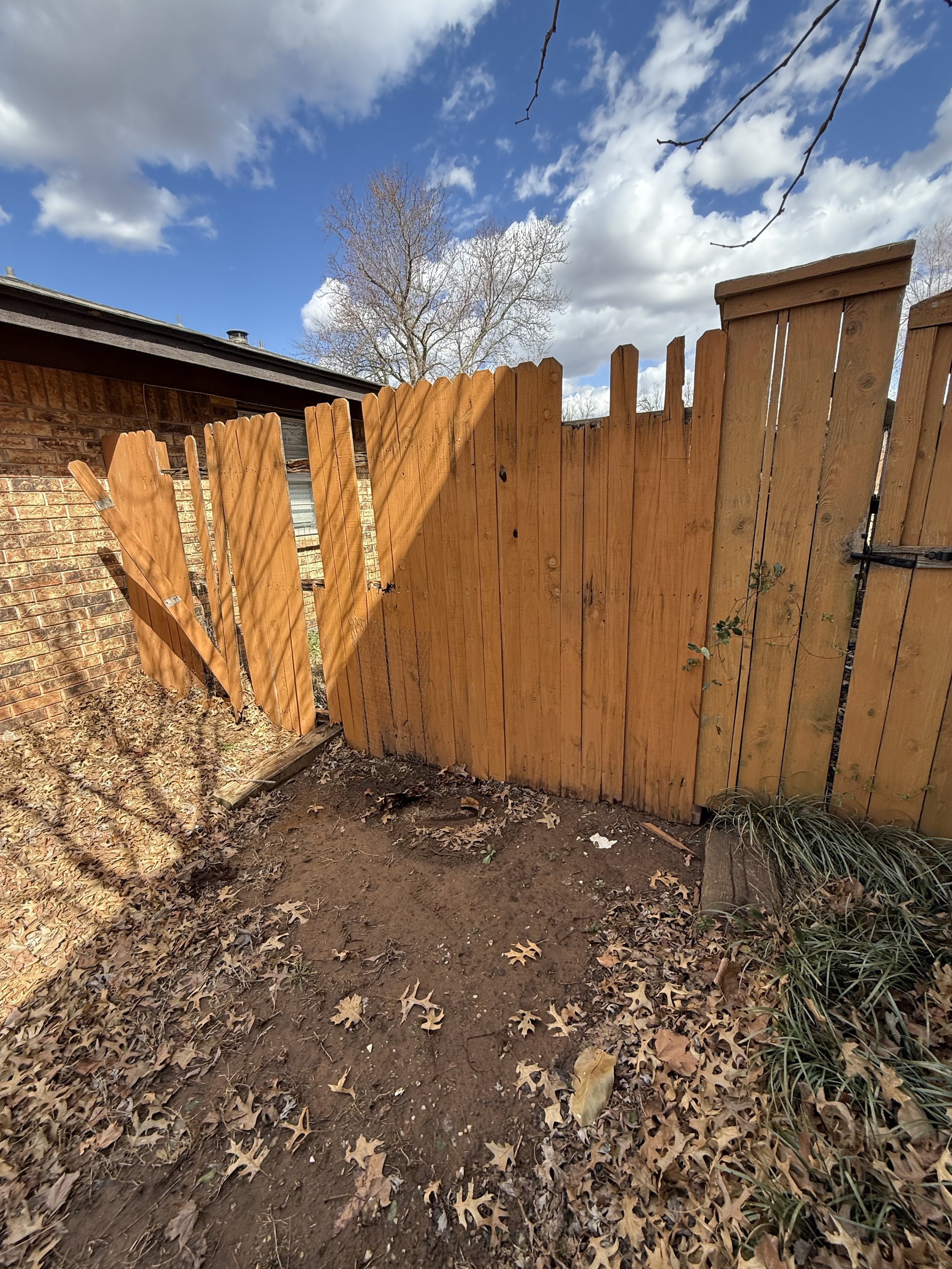 Broken wooden fence with missing panels, dirt ground with fallen leaves, and a brick building in the background under a cloudy sky.