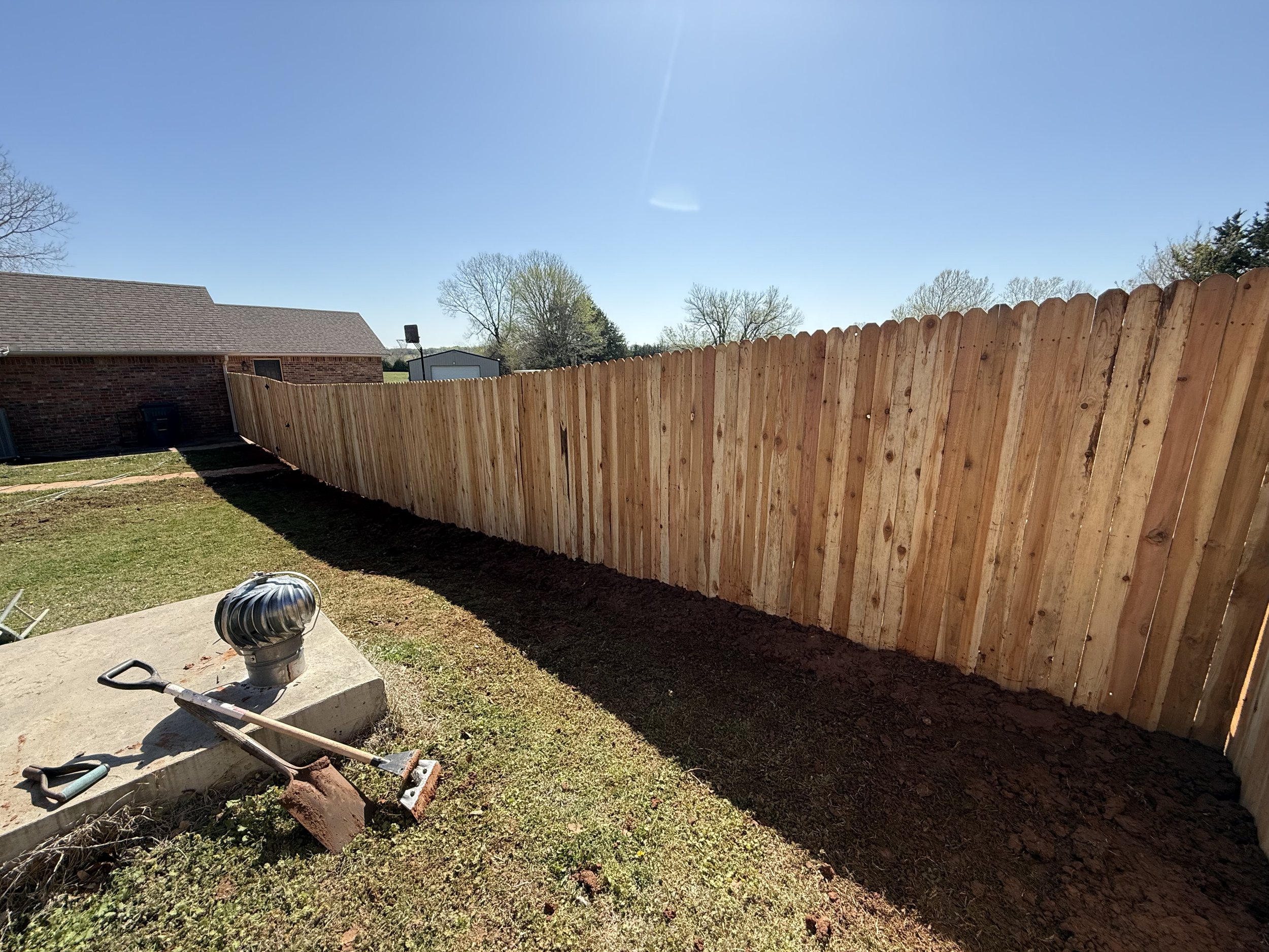 Wooden fence enclosing a backyard with garden tools on the grass, under a clear blue sky.