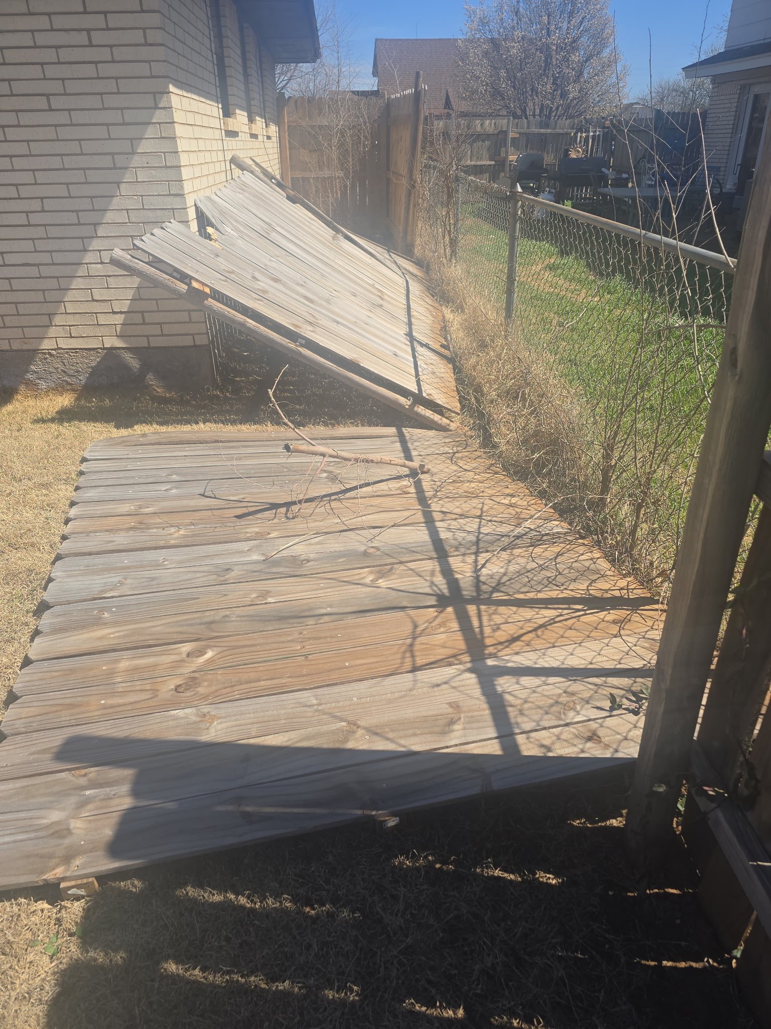 Wooden fence panels fallen and leaning against a brick wall next to a chain-link fence in a backyard.