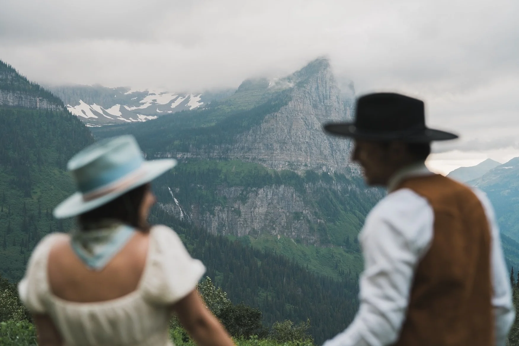 A man and a woman dressed in vintage clothing with wide-brimmed hats, standing outdoors with mountains and clouds in the background in Glacier National Park