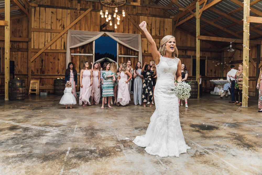 A bride in a white dress prepares to throw her bouquet in a rustic barn setting, with a group of women and a child gathered in anticipation.