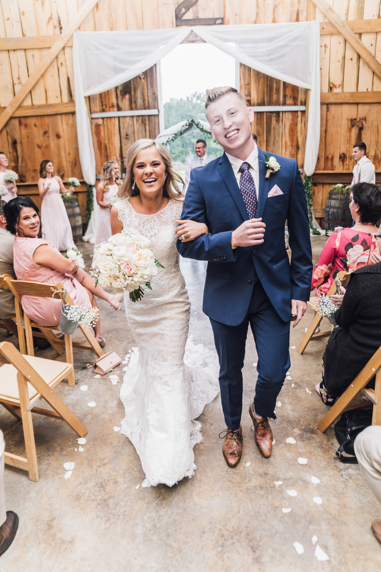 A bride and groom walking down the aisle at a rustic indoor wedding venue, smiling and holding hands, surrounded by guests seated in wooden chairs. The bride is wearing a white lace dress and holding a bouquet. The groom is in a navy suit with a flor
