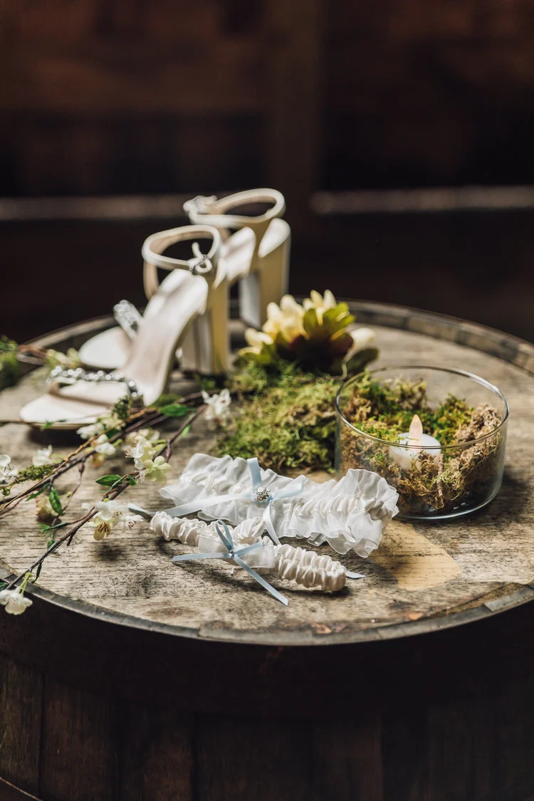 Wedding accessories on rustic wooden barrel including white high heels, lace garters with blue ribbons, floral decorations, and a candle in a glass bowl with moss.