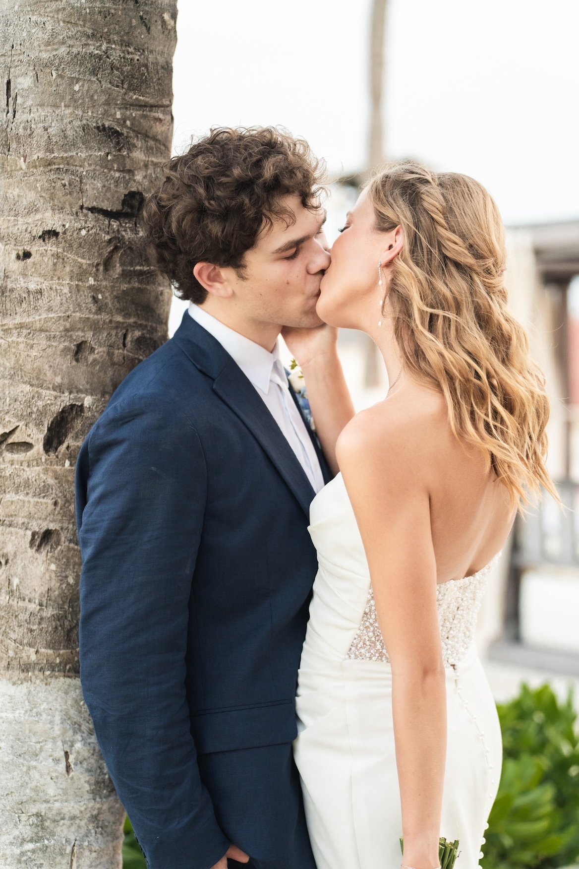 A man and woman sharing a kiss outdoors, with the woman dressed in a white strapless wedding gown and the man in a dark blue suit, next to a tree in Cancun, Mexico.