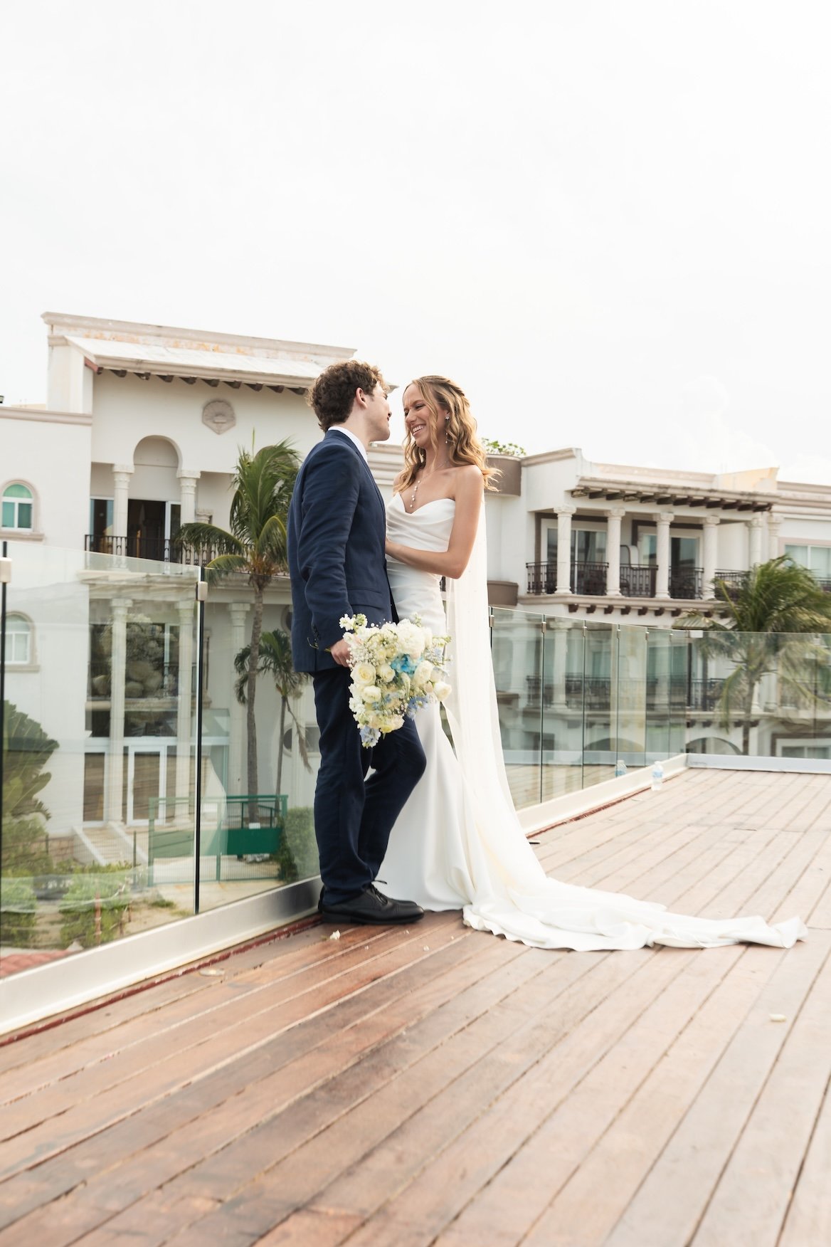 A bride and groom stand close together on a rooftop, facing each other and smiling. The bride holds a bouquet of white and blue flowers. The setting includes modern white buildings in Cancun, Mexico.