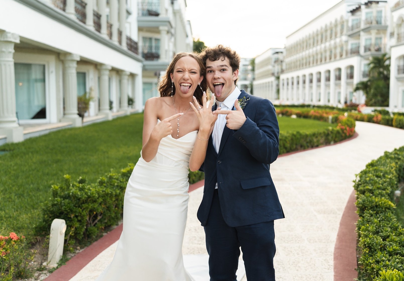 A happy couple in wedding attire making silly faces and pointing at each other outdoors near a large white building with balconies and lush green landscaping  in Cancun, Mexico.