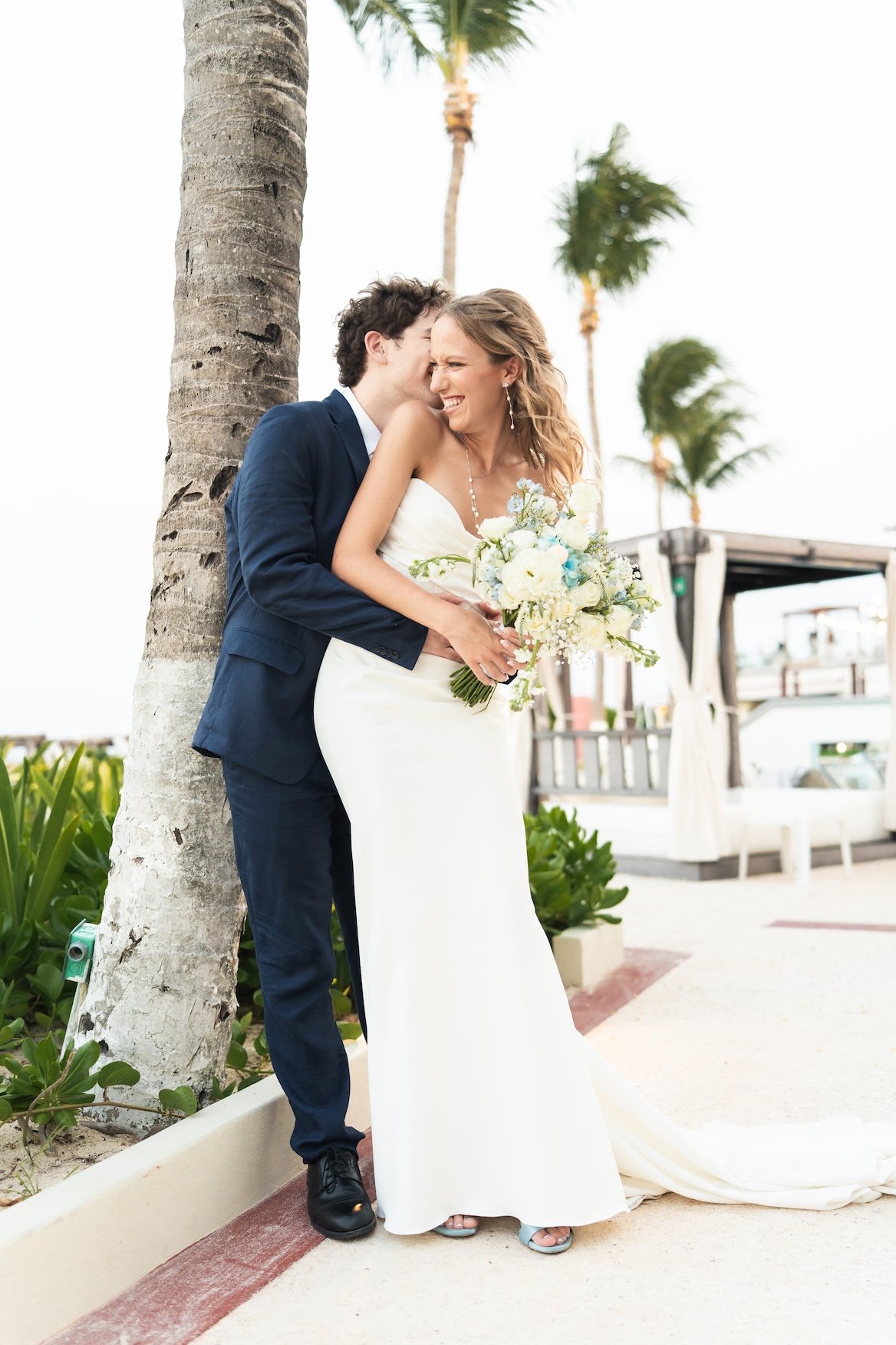 A newlywed couple in wedding attire sharing a joyful moment outdoors near palm trees, with the groom whispering in the bride's ear and the bride holding a bouquet of white flowers in Cancun, Mexico.