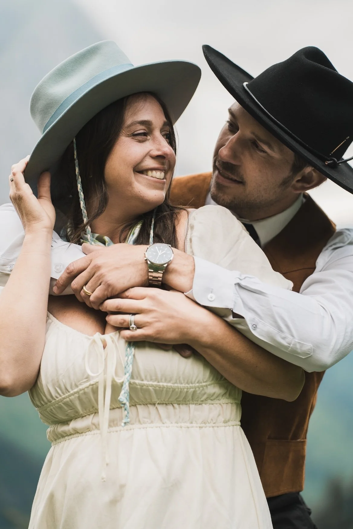 A happy couple wearing cowboy hats smiling and embracing in Glacier National Park, with the woman wearing a beige dress and the man in a white shirt and brown vest.