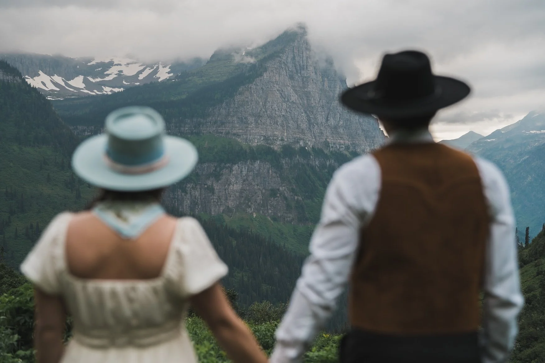 A couple dressed in vintage clothing holds hands while looking at a misty mountain range with lush green forests in Glacier National Park.