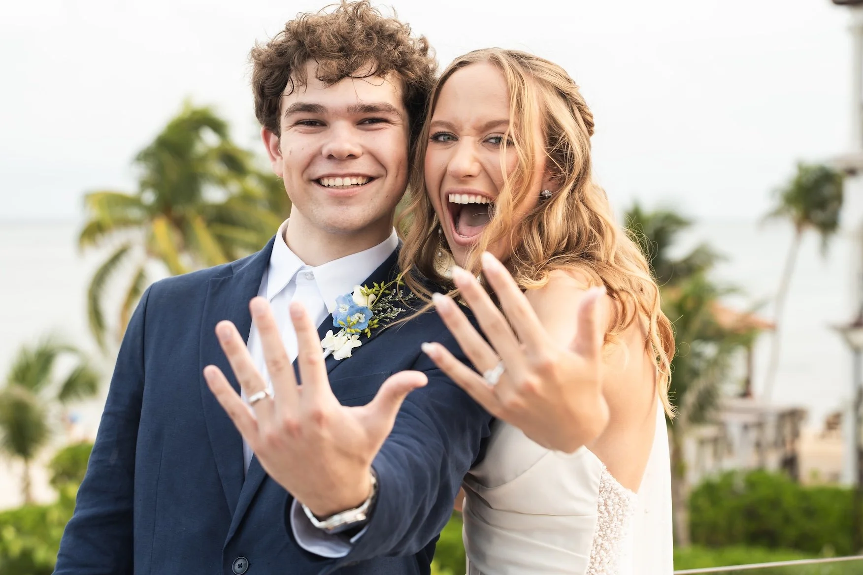 a smiling young man and woman celebrating a wedding, showing their rings with a happy expression, outdoors with palm trees in the background in Cancun, Mexico.