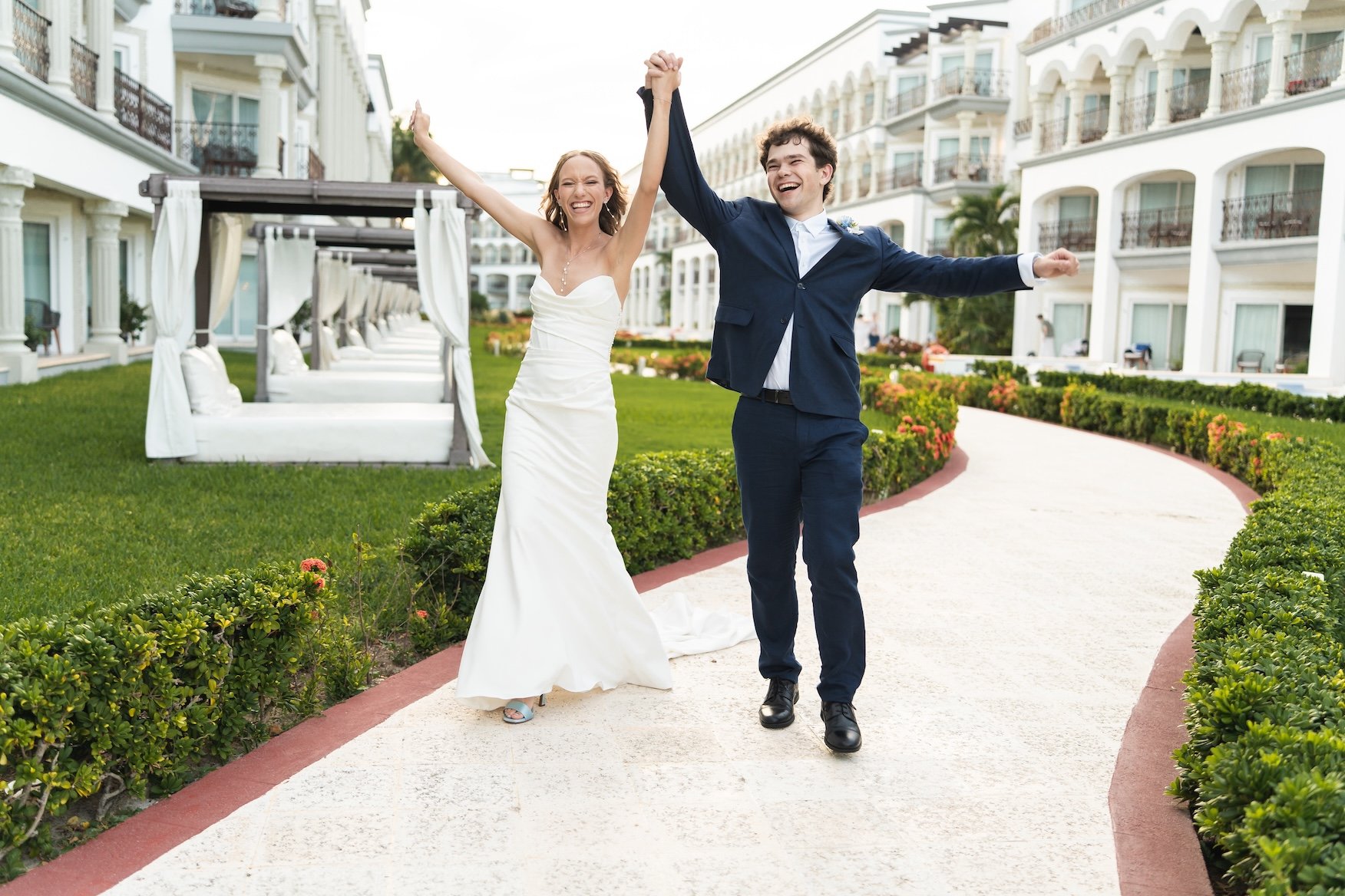 A newlywed couple celebrates on a pathway outside a large white hotel or resort, smiling and holding hands in the air  in Cancun, Mexico.