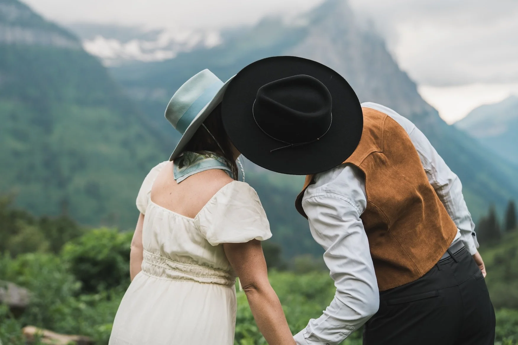 A couple leaning close together outdoors with mountains and greenery in the background, wearing hats and vintage-style clothing in Glacier National Park