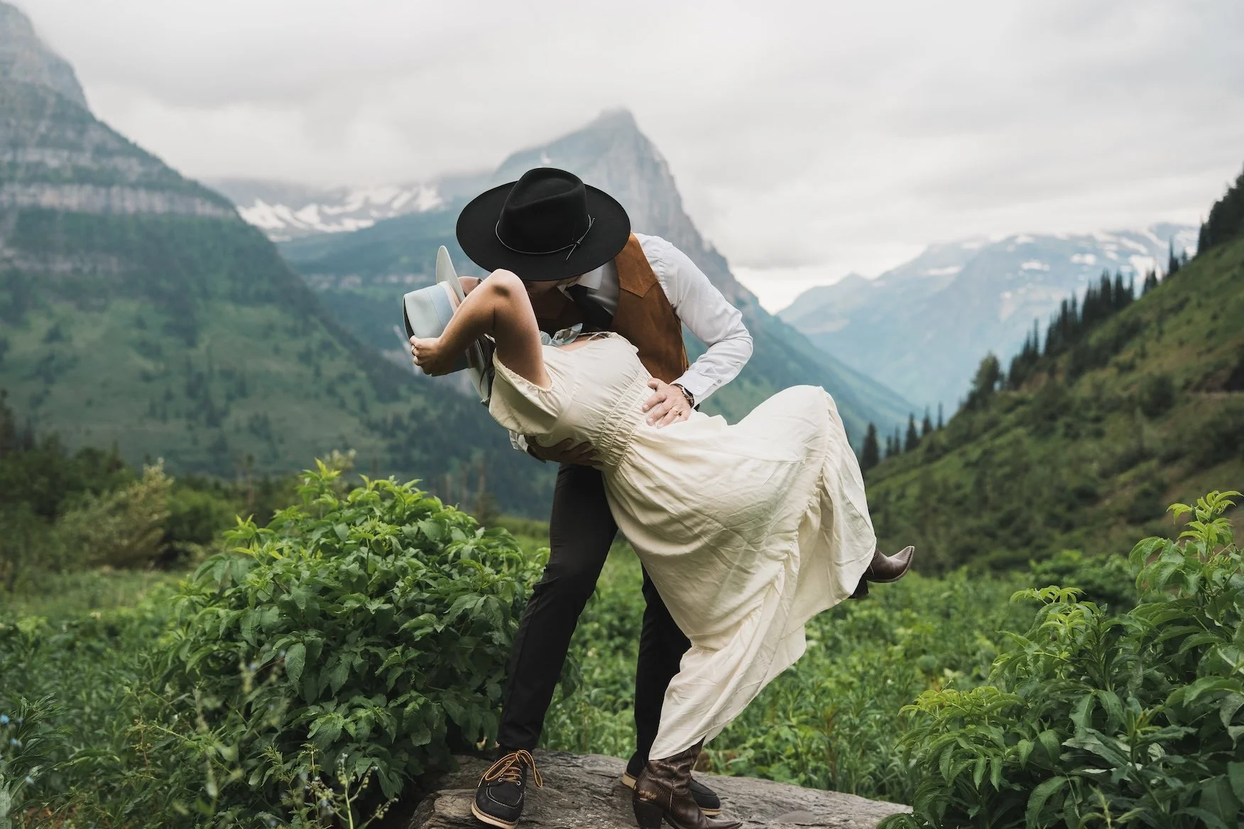 A man in vintage clothing and a woman in a cream dress and hat are dancing outdoors on a rock in Glacier National Park