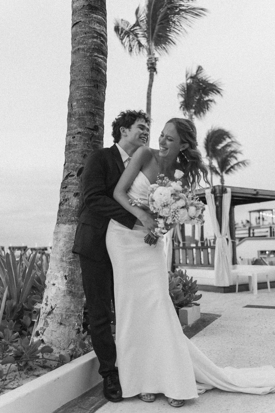A couple in wedding attire, smiling and embracing outdoors near palm trees, with a bouquet of flowers, on a beachside in Cancun.