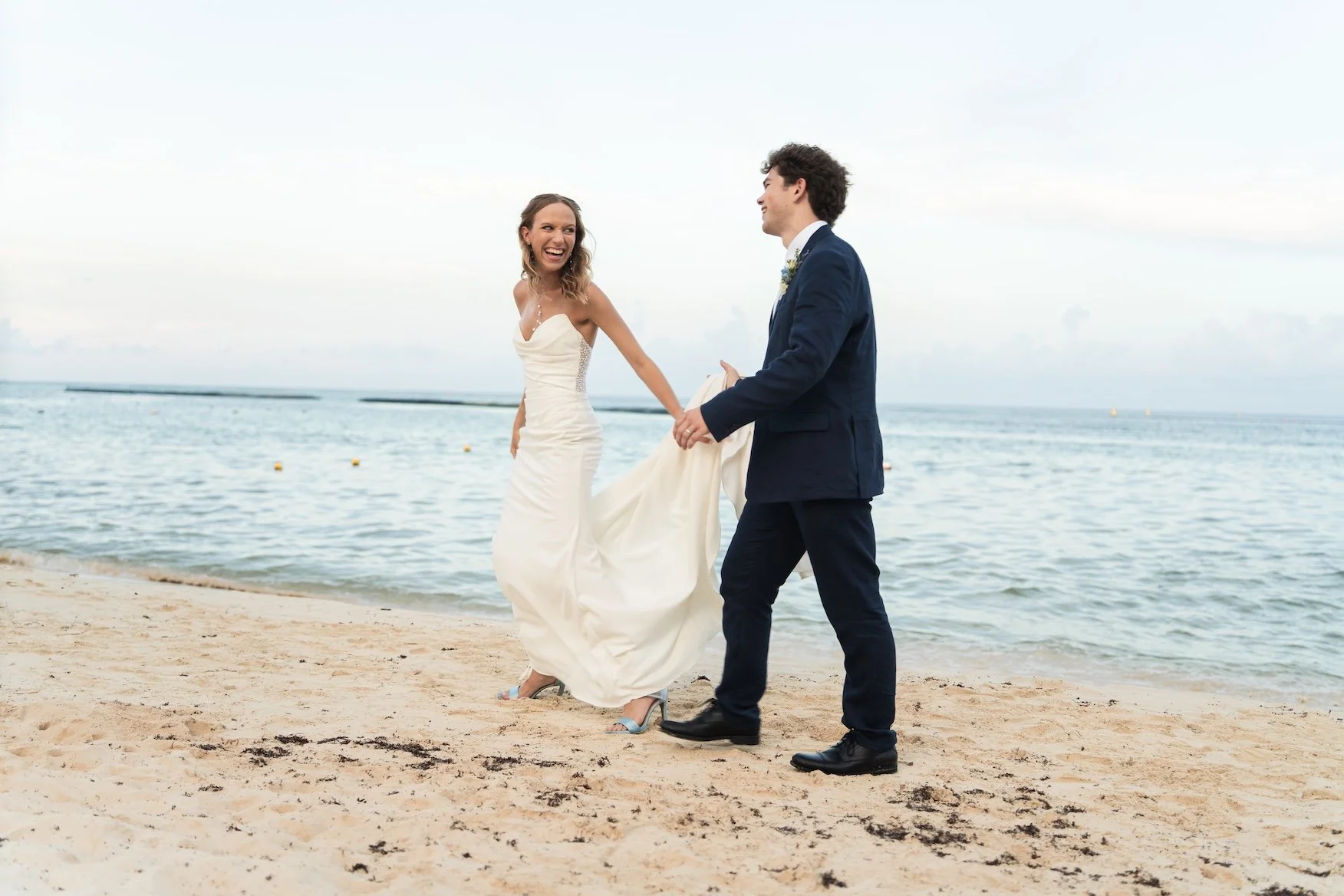 A couple, dressed in wedding attire, on a beach, holding hands and smiling at each other with the ocean in the background in Cancun, Mexico.