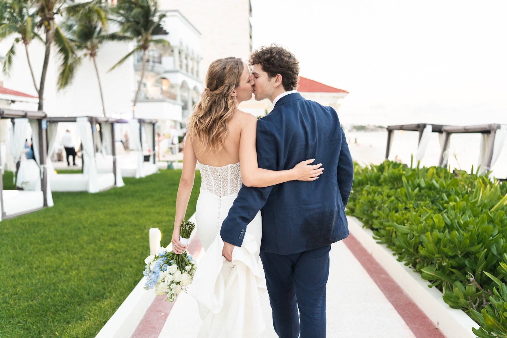 A bride and groom sharing a kiss outdoors, with the bride holding a bouquet of flowers and wearing a strapless wedding gown, and the groom in a dark suit, on a pathway lined with greenery and beach cabanas in the background in Cancun, Mexico.