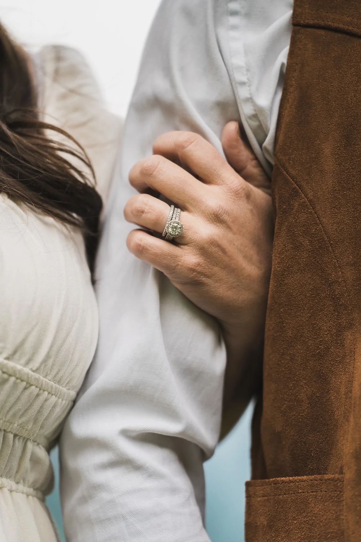 Close-up of a woman's hand with an engagement ring on her ring finger, resting on a man's chest, who is wearing a white shirt and brown jacket in Glacier National Park
