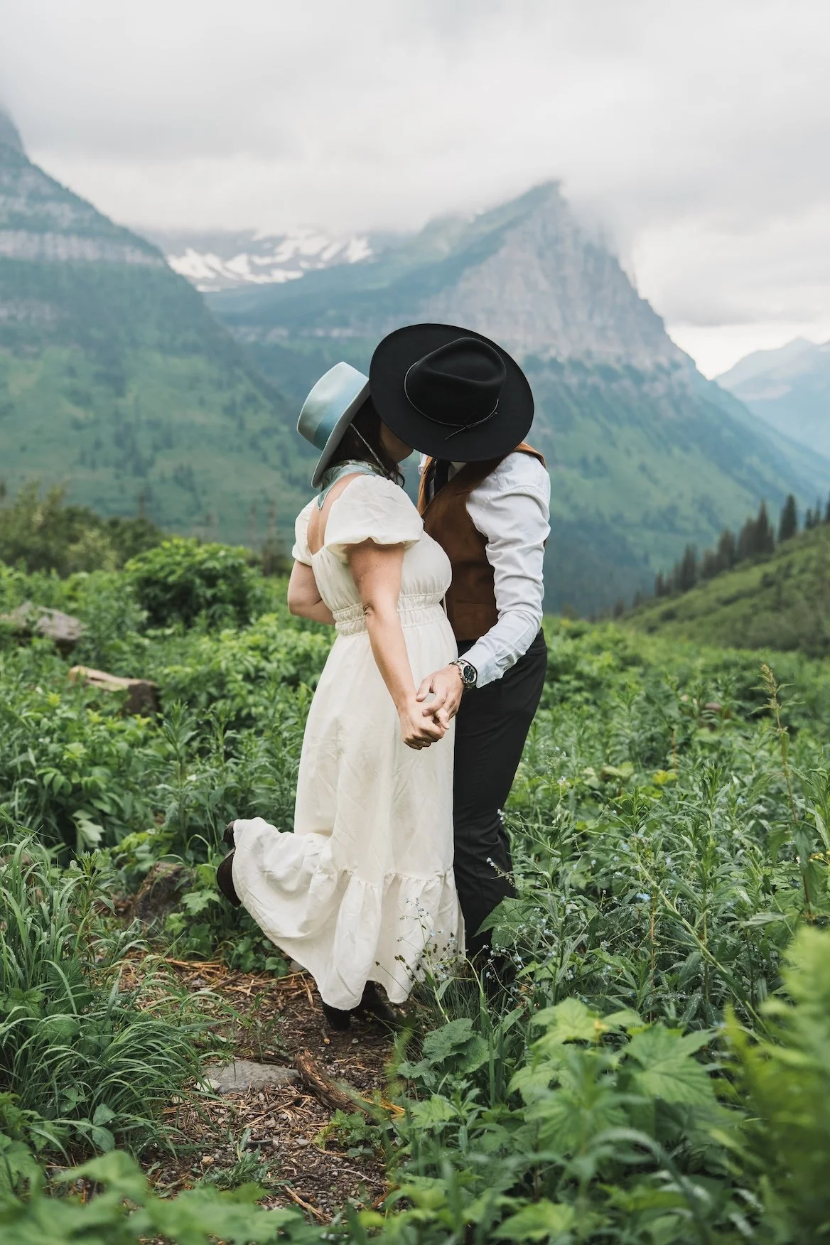 Couple holding hands and kissing in a green mountain field, wearing vintage-style clothing and black hats in Glacier National Park