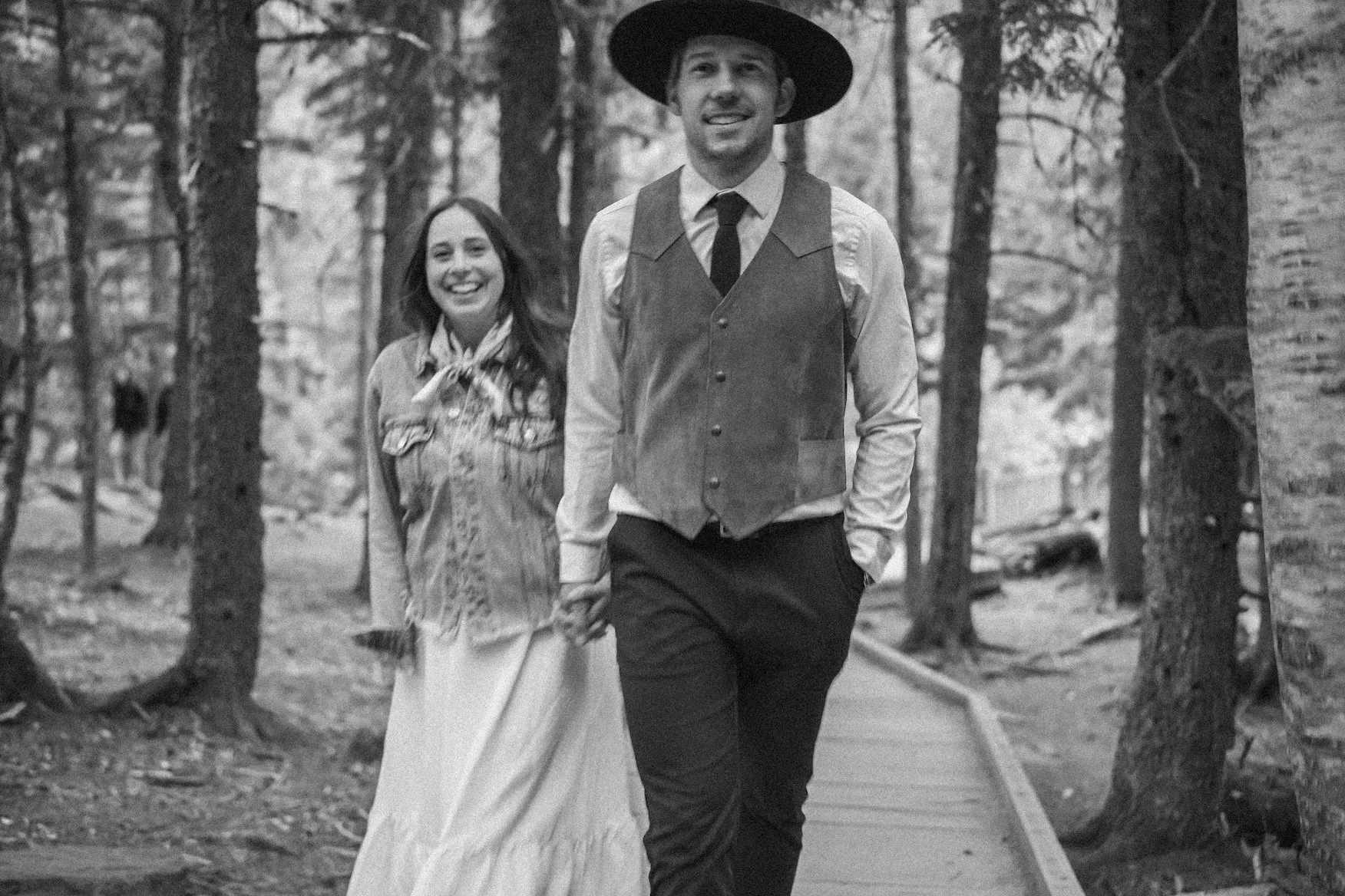 A man and woman walking hand in hand on a forest path in Glacier National Park, smiling and enjoying each other's company, in black and white.