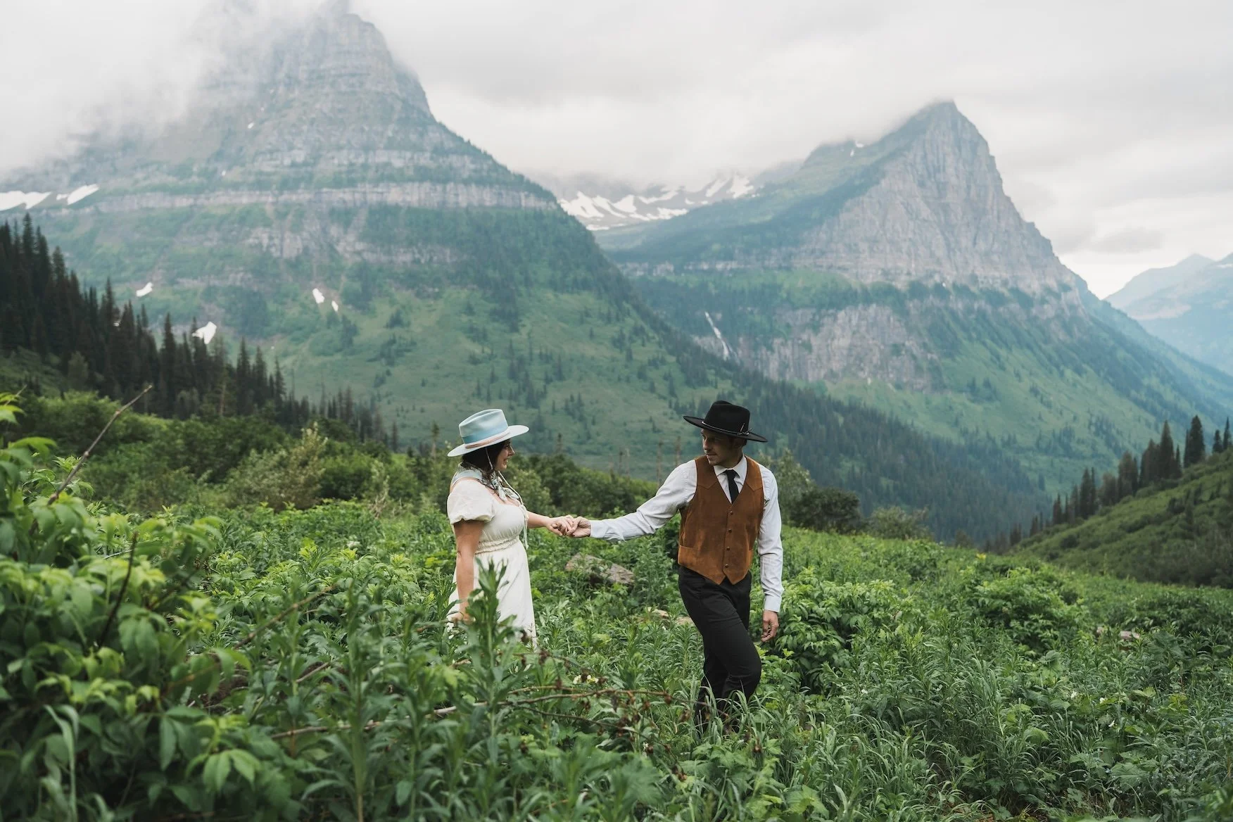 A man and woman dressed in vintage clothing holding hands in a green field in Glacier National Park.