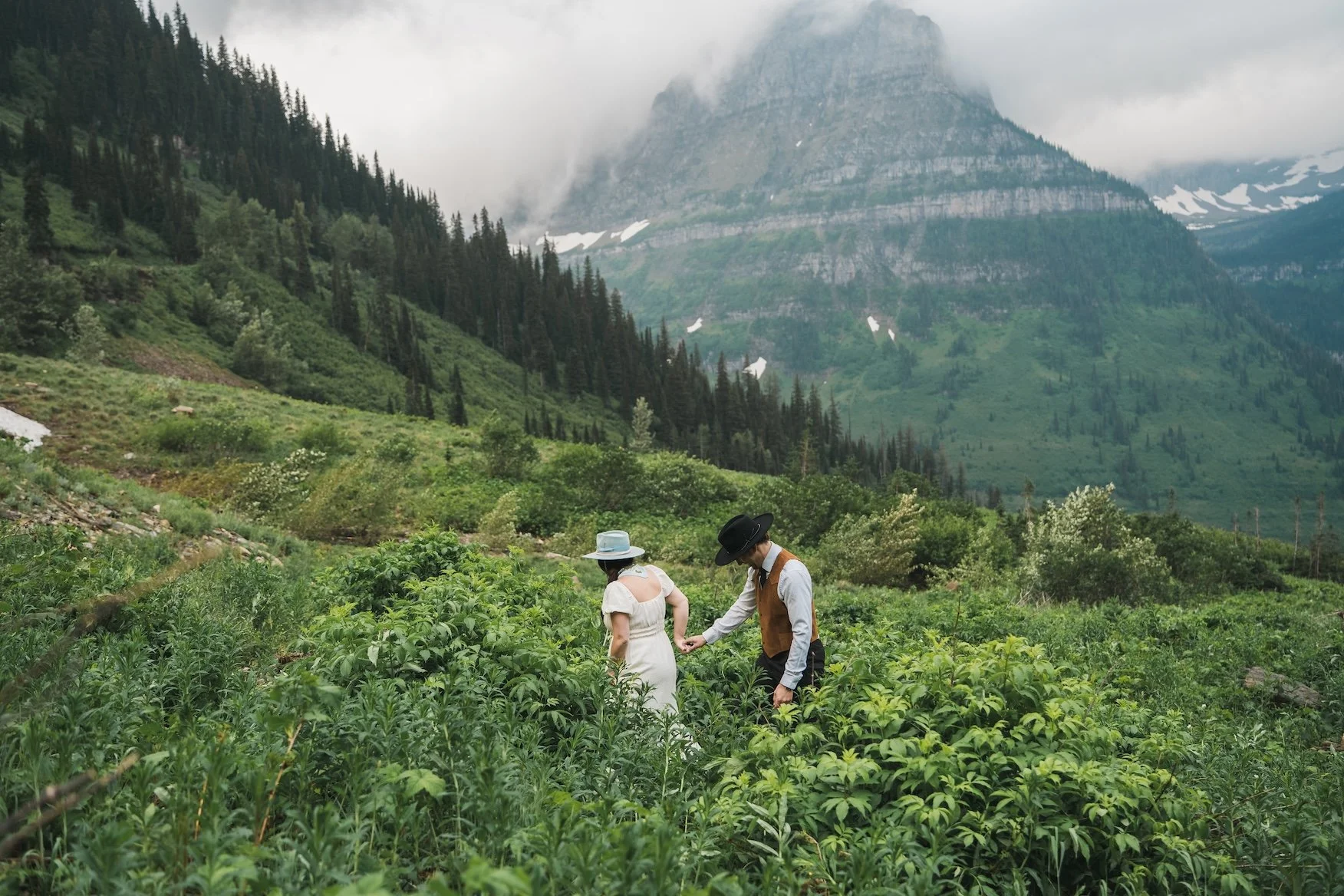 A man and woman in vintage clothing exploring a lush green field with tall mountains and cloudy sky in the background in Glacier National Park
