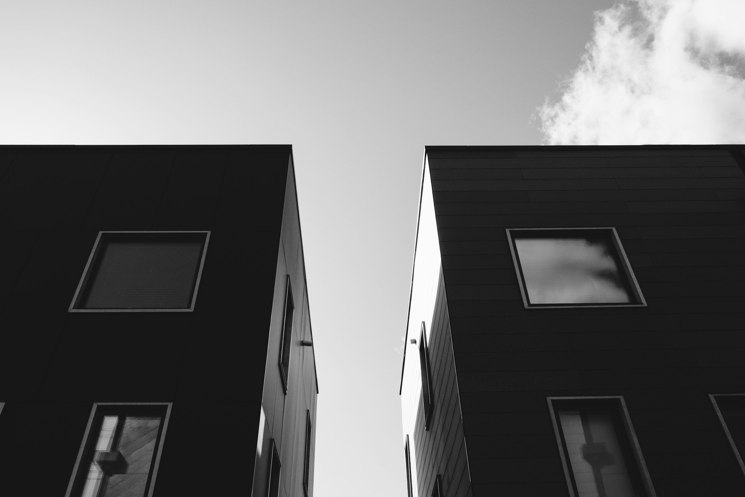Black and white photograph of two modern buildings with rectangular windows, viewed from below, against a partly cloudy sky.