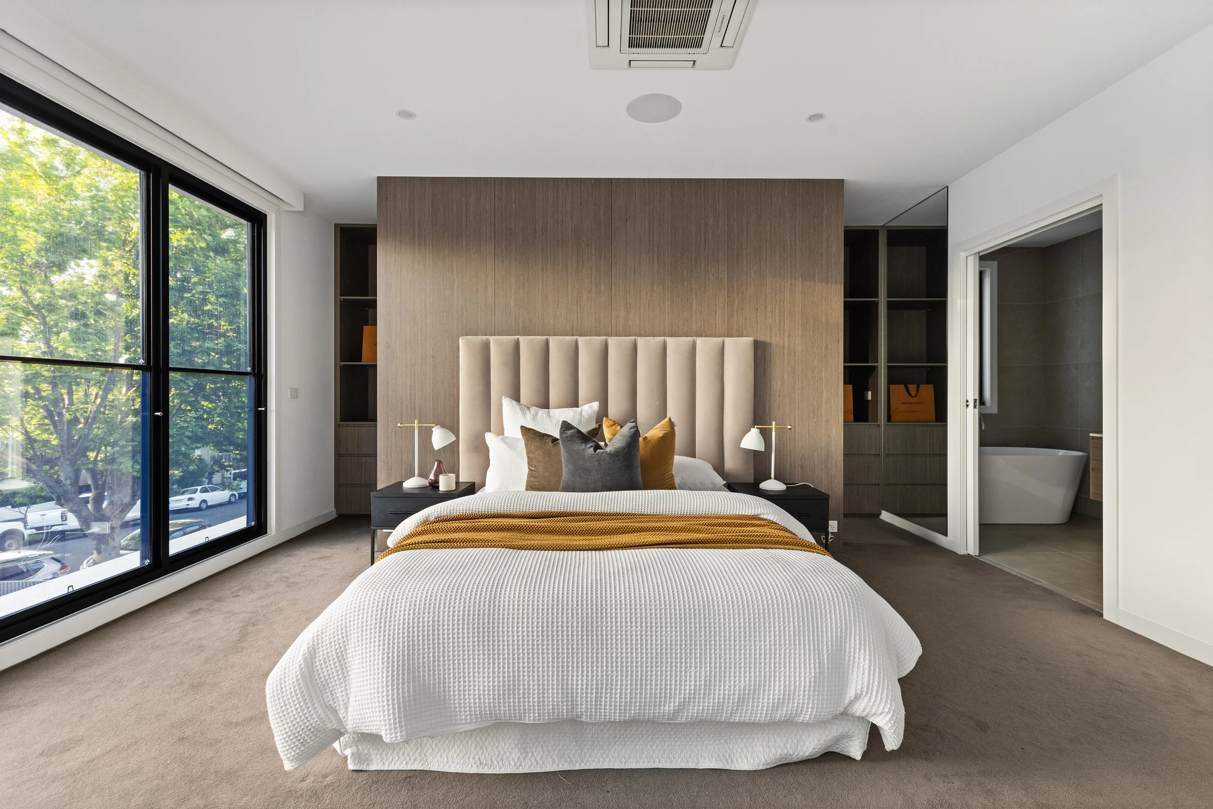 Modern bedroom with a large bed featuring a beige upholstered headboard, white bedding, various colored pillows, and a mustard throw blanket. Two bedside tables with white lamps flank the bed. Large windows on the left offer a view of trees outside. An open door on the right reveals a bathroom with a white bathtub and beige tiles.