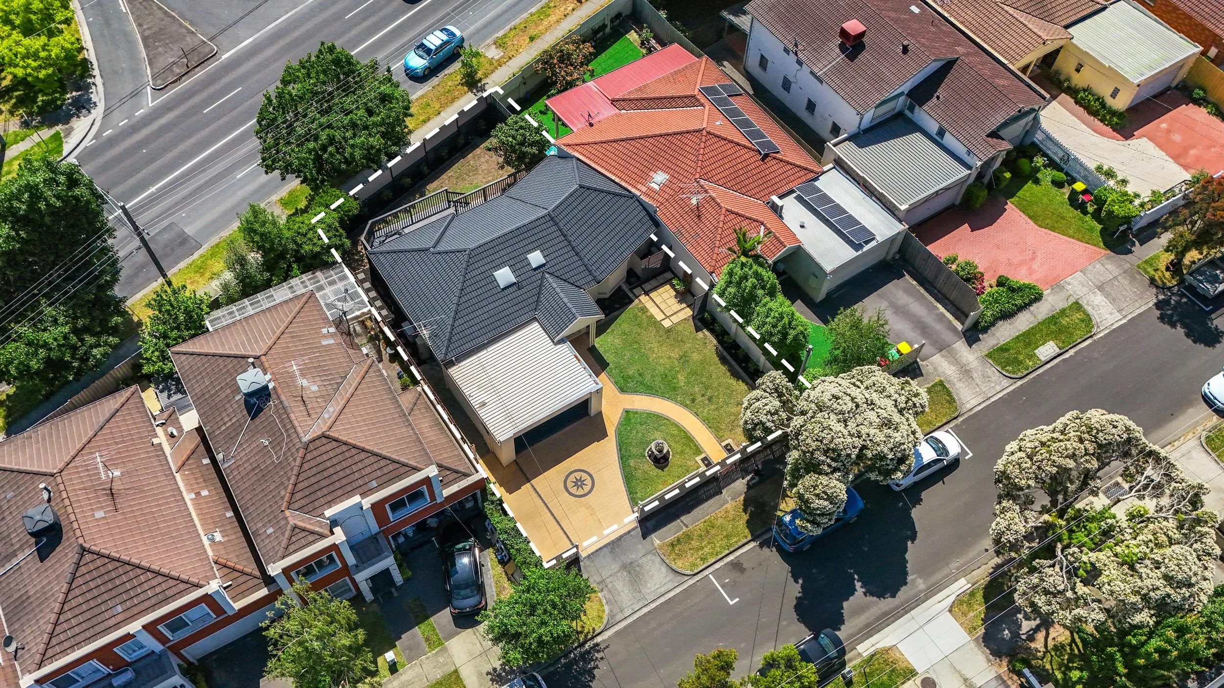 Aerial view of a residential neighborhood with several single-family houses, trees, parked cars, and a main road. The houses have different colored roofs, some with solar panels, and fenced yards.