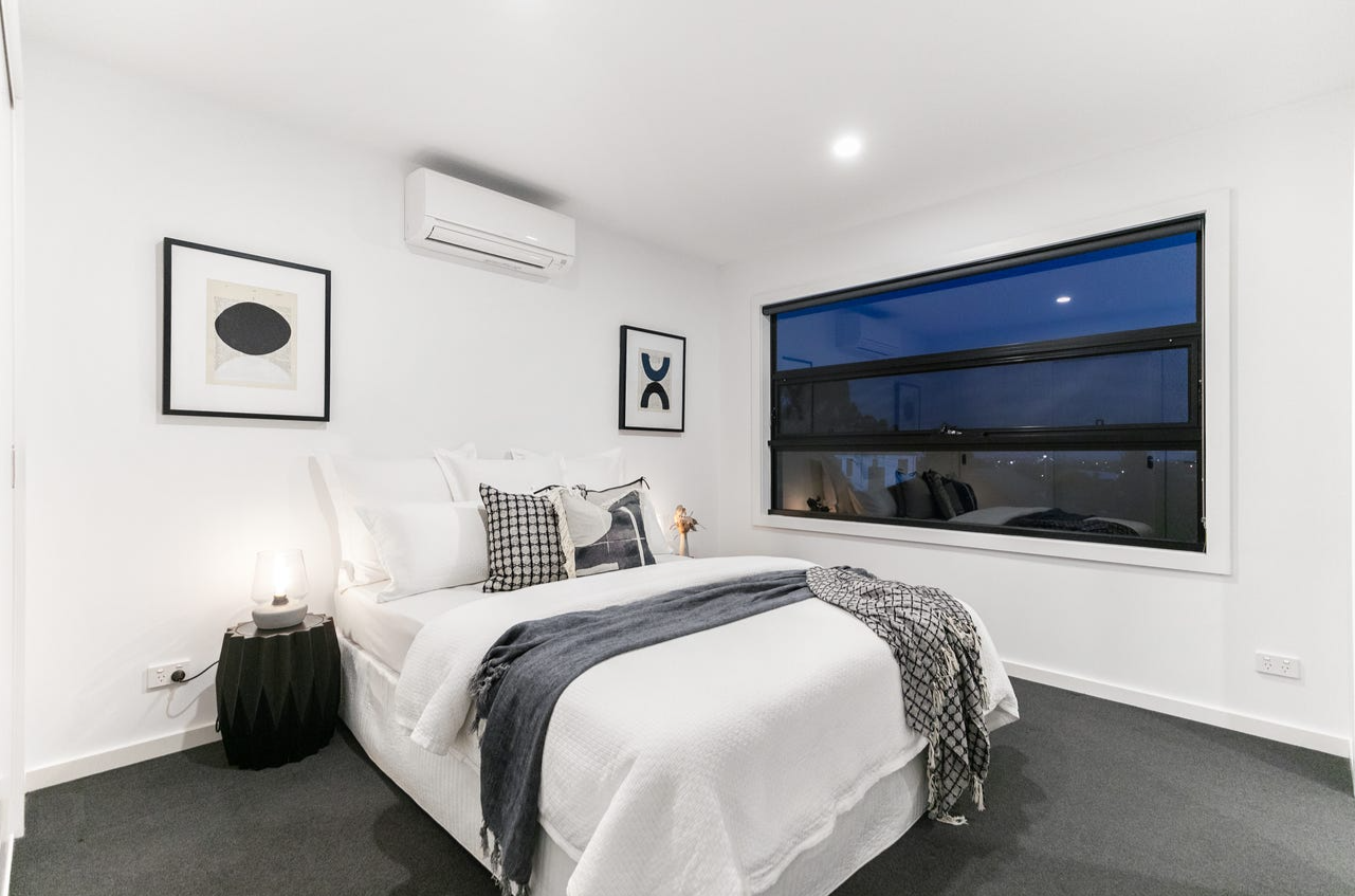 A modern bedroom with white walls, a large window at night, a decorated bed with throw pillows and blankets, framed abstract art on the walls, an air conditioning unit, and a black side table with a lamp.