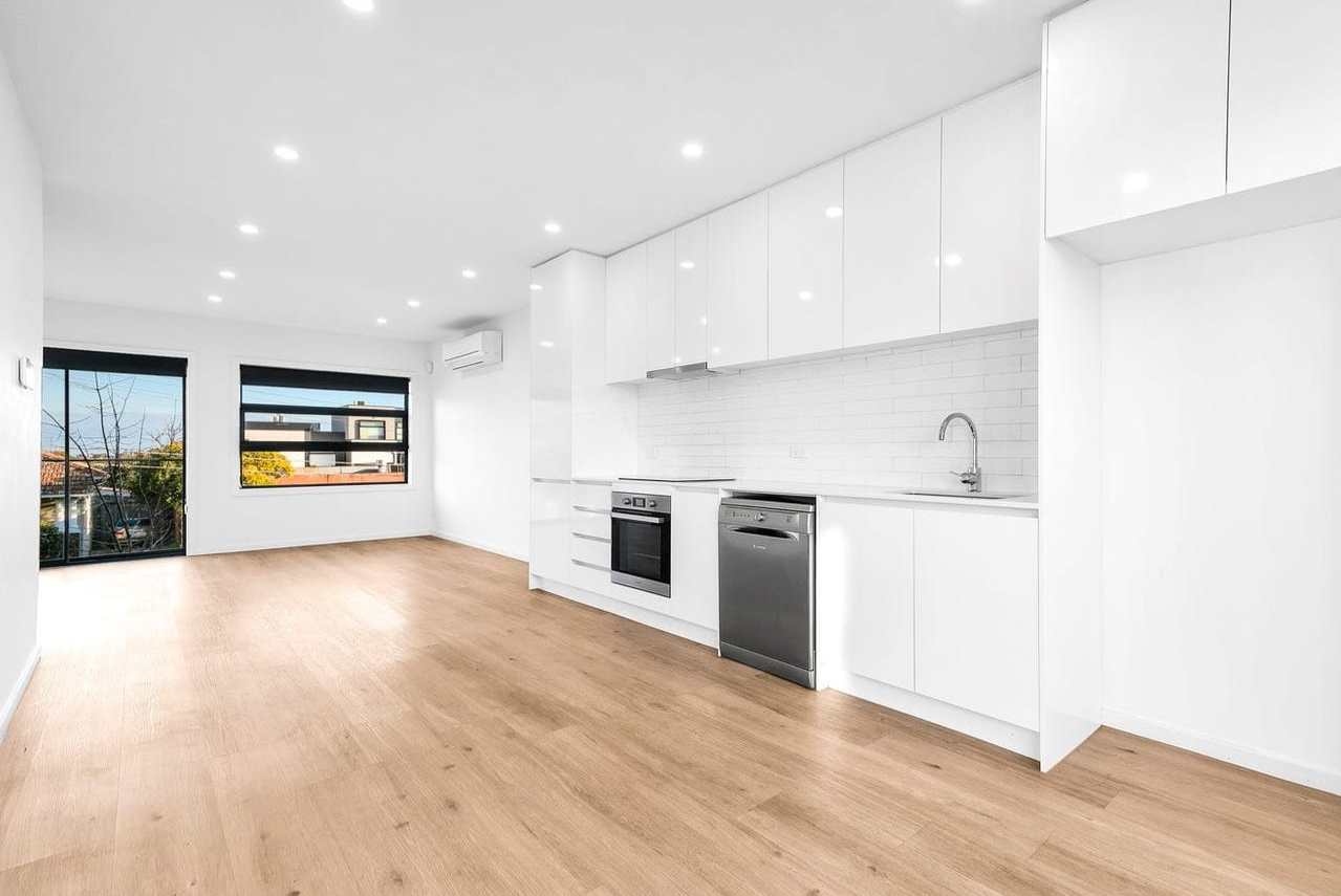 Empty modern kitchen and living room with wooden floors, white cabinets, white walls, and large windows showing trees and buildings outside.
