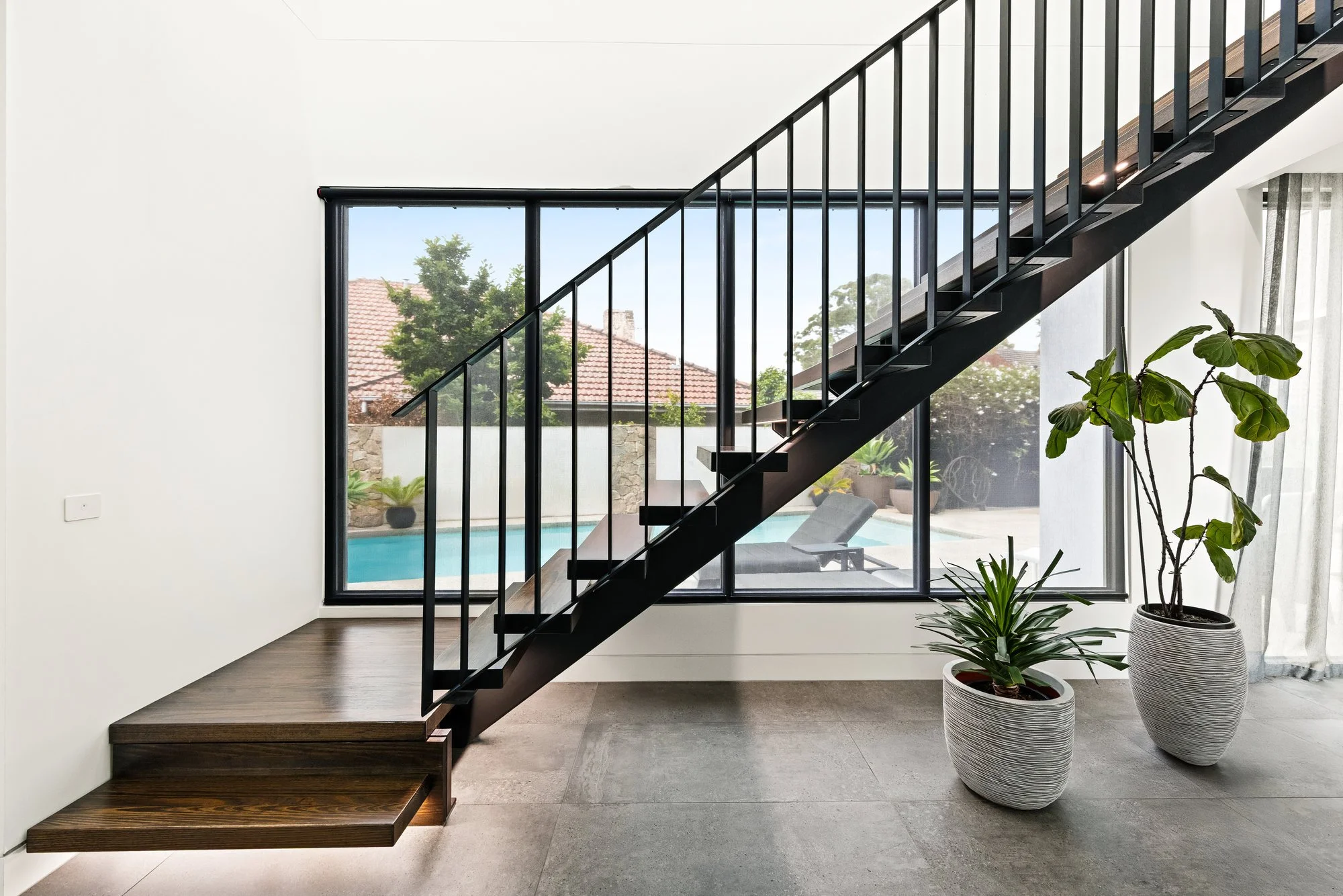 Modern interior with black metal staircase, large glass window showing outdoor pool and patio, two potted plants, white walls and tiled floor.